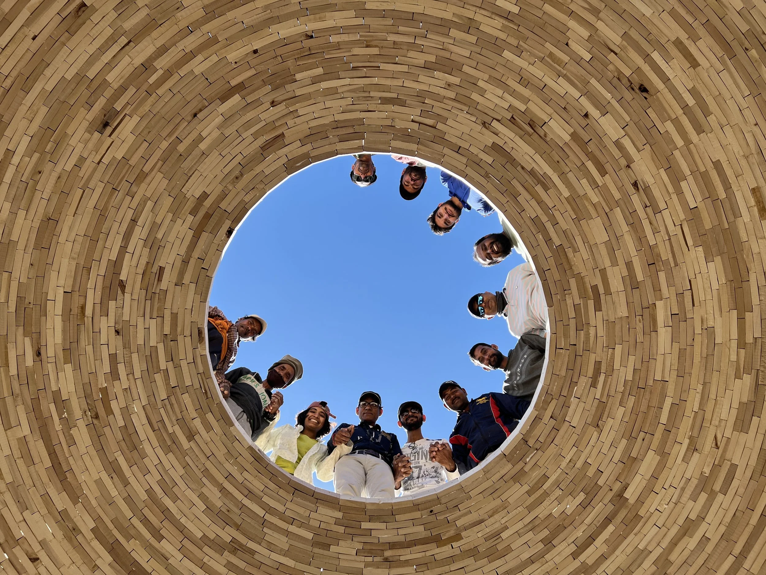 View looking up into a circular wooden structure with people standing around the edge, looking down at the camera, against a clear blue sky.