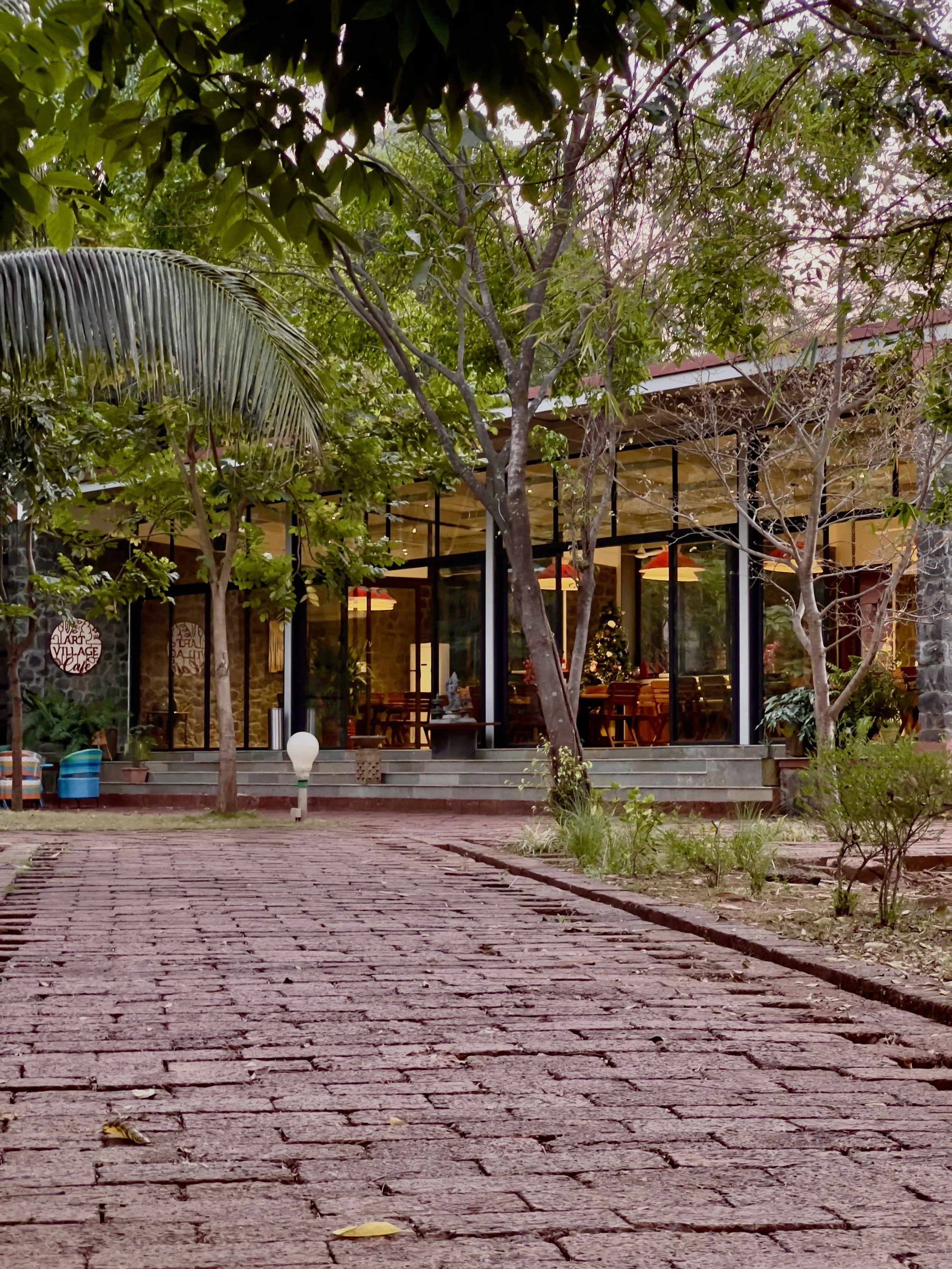 A brick pathway leading to a glass-fronted cafe or restaurant surrounded by trees and greenery.