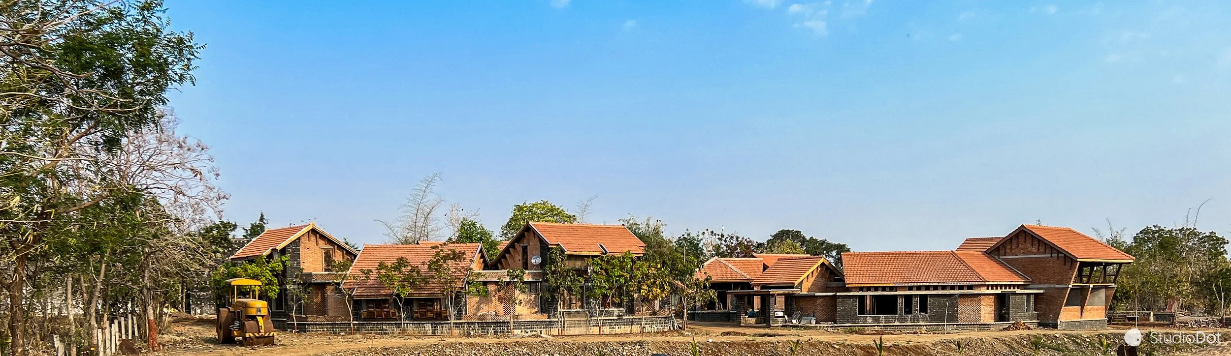 Under construction houses with a yellow construction vehicle in front, trees, and a clear blue sky.