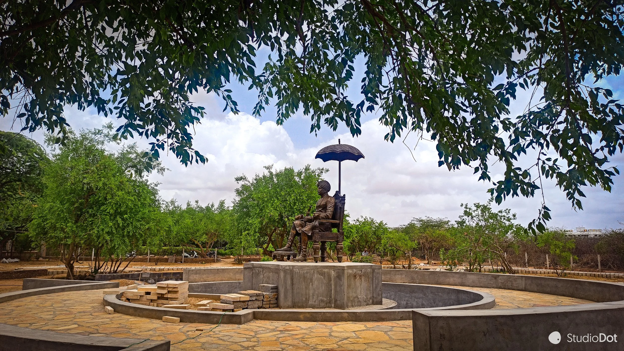 Statue of a woman sitting on a chair holding an umbrella in a park with trees and cloudy sky in the background.