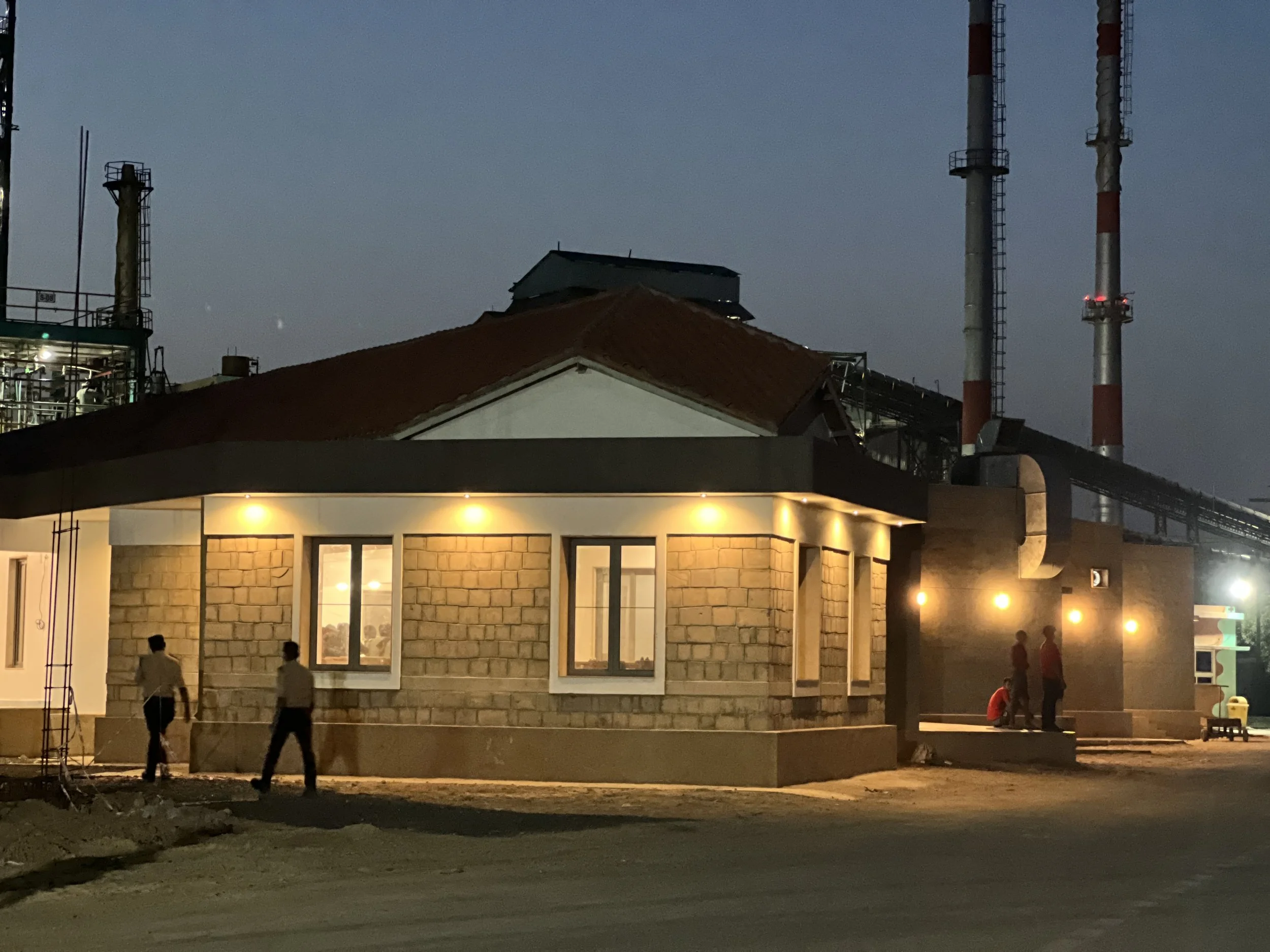 A building with lit exterior lights at dusk, featuring windows and a brick facade, with four people standing outside and industrial structures and smokestacks in the background.