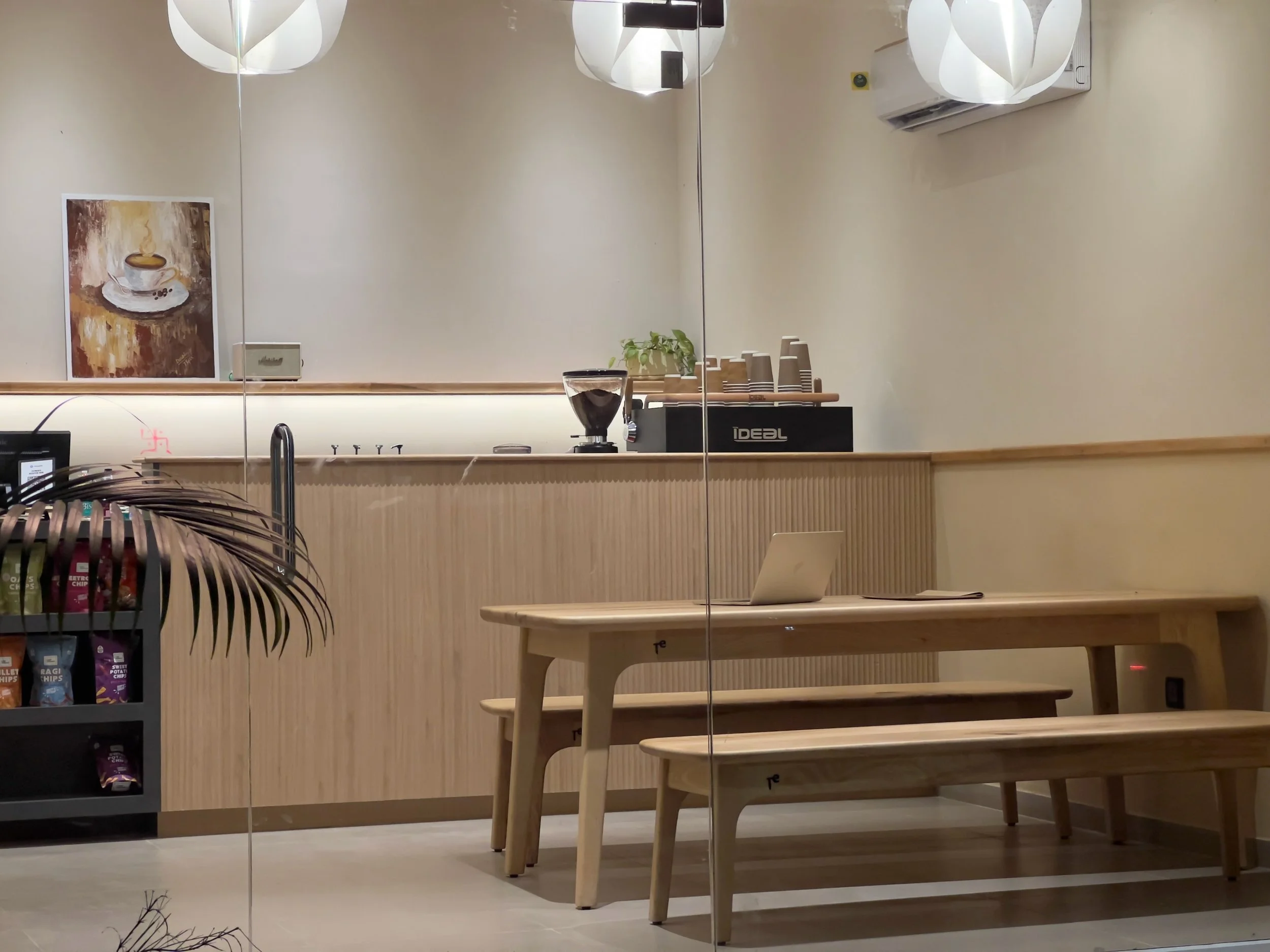 View of a minimalist coffee shop counter with a coffee grinder, cups, and a small plant; a bench and table are in the foreground, and a wall-mounted air conditioning unit is visible.