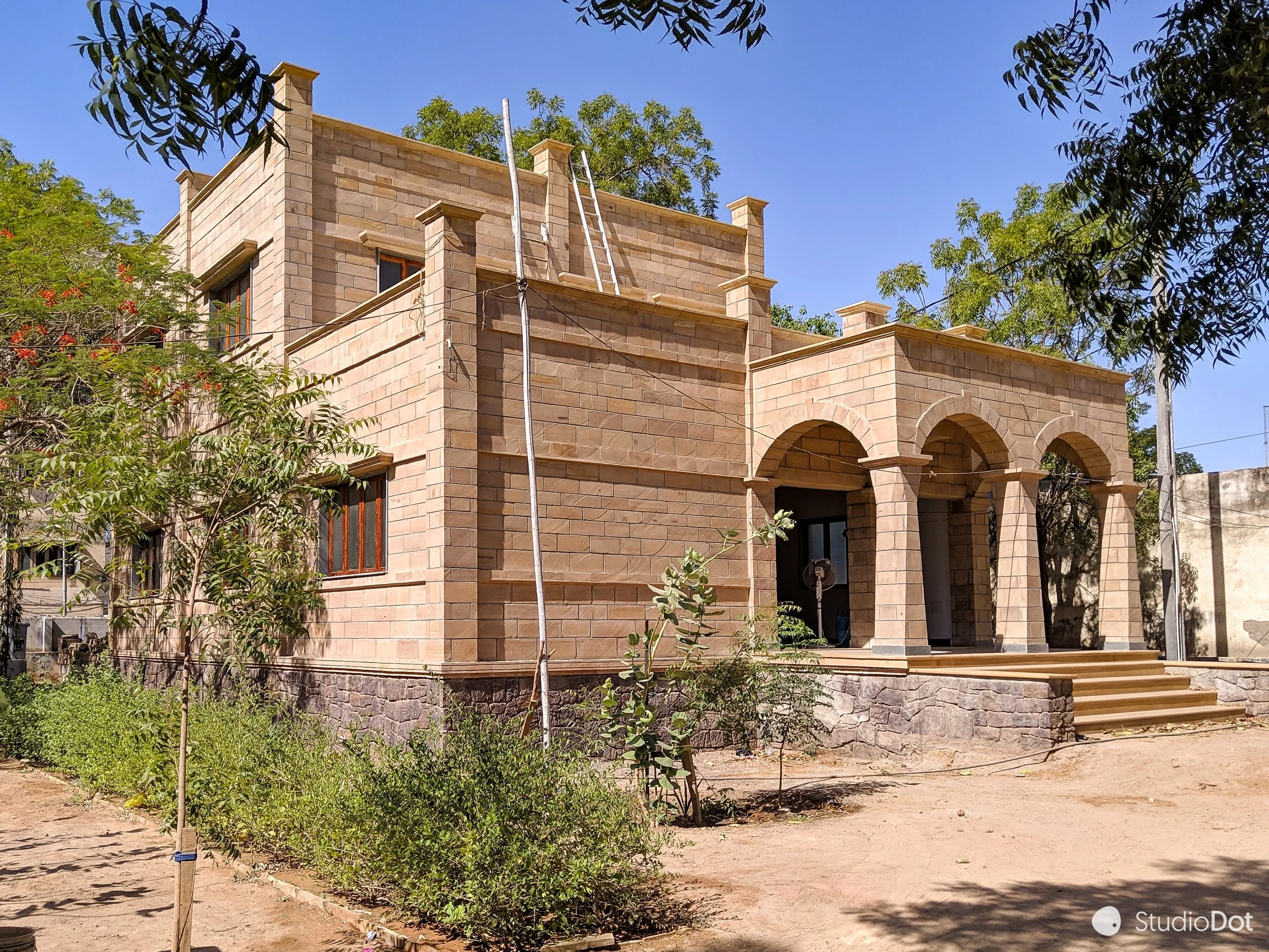 A large stone building under construction with arched doorways and a staircase, surrounded by trees and dirt ground.