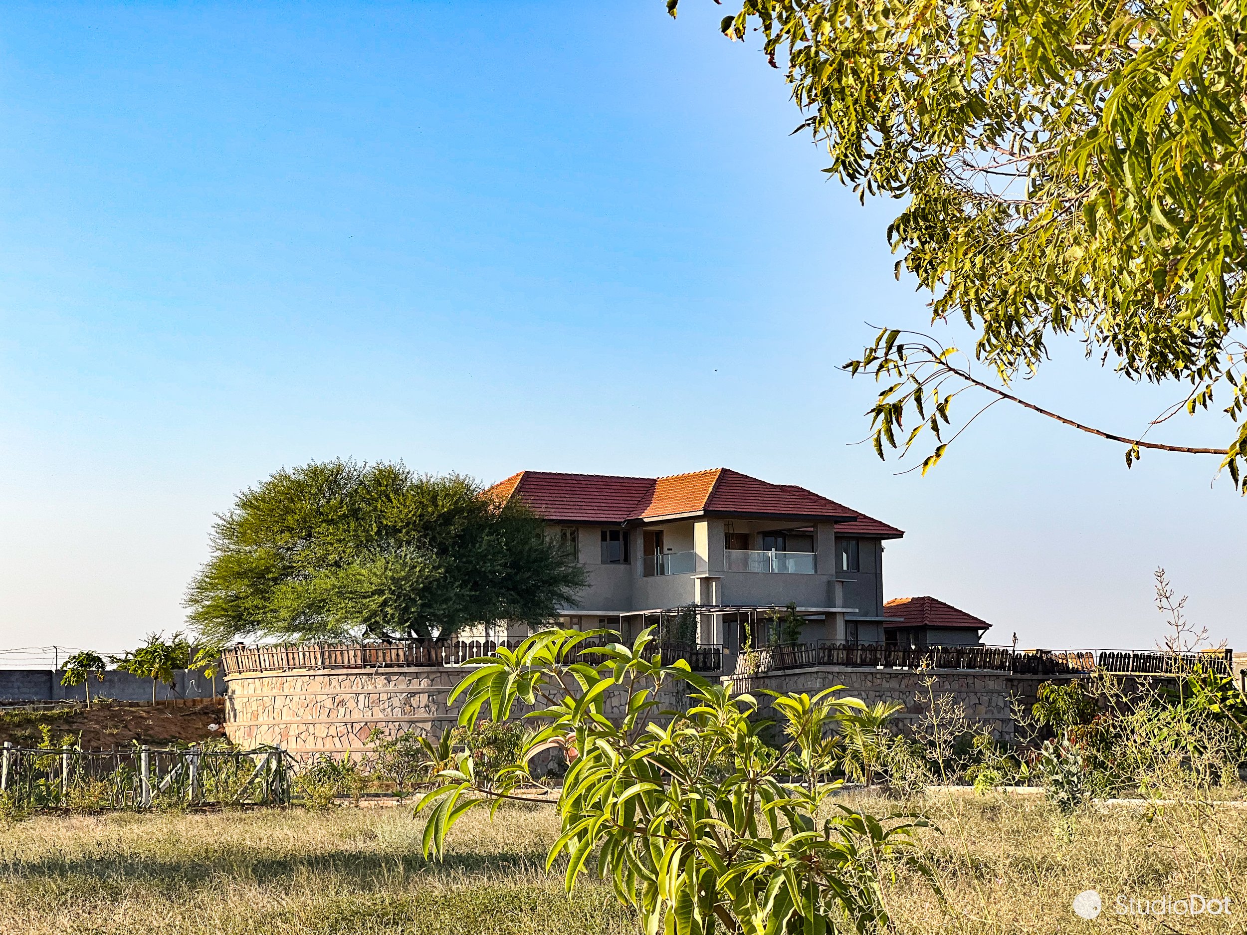 A two-story house with a red-tiled roof and balconies, situated on a raised stone platform, surrounded by trees and bushes in a grassy area, under a clear blue sky.