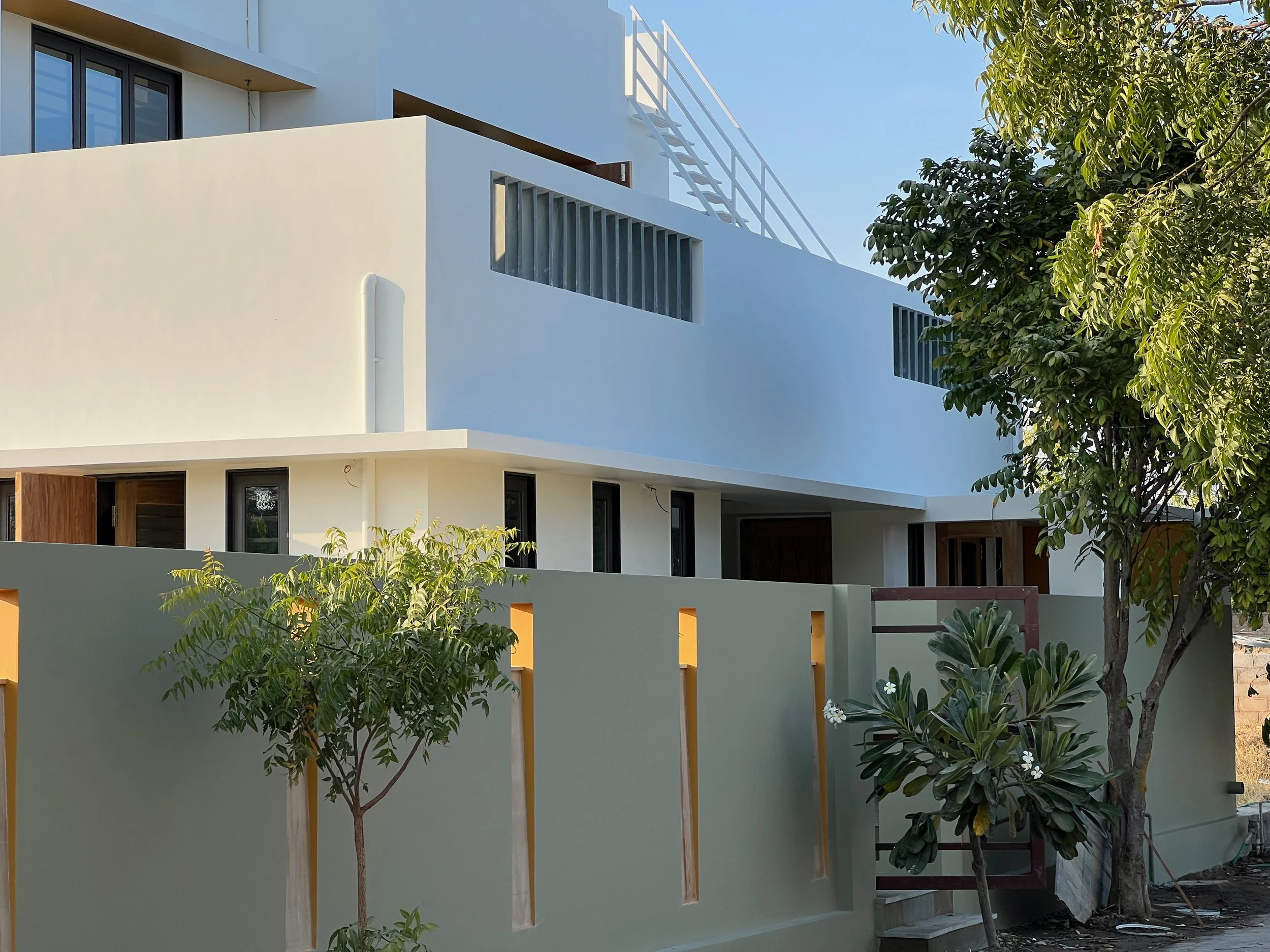 Modern white multi-story residential building with a gray boundary wall, trees, and plants in the foreground, under a clear blue sky.