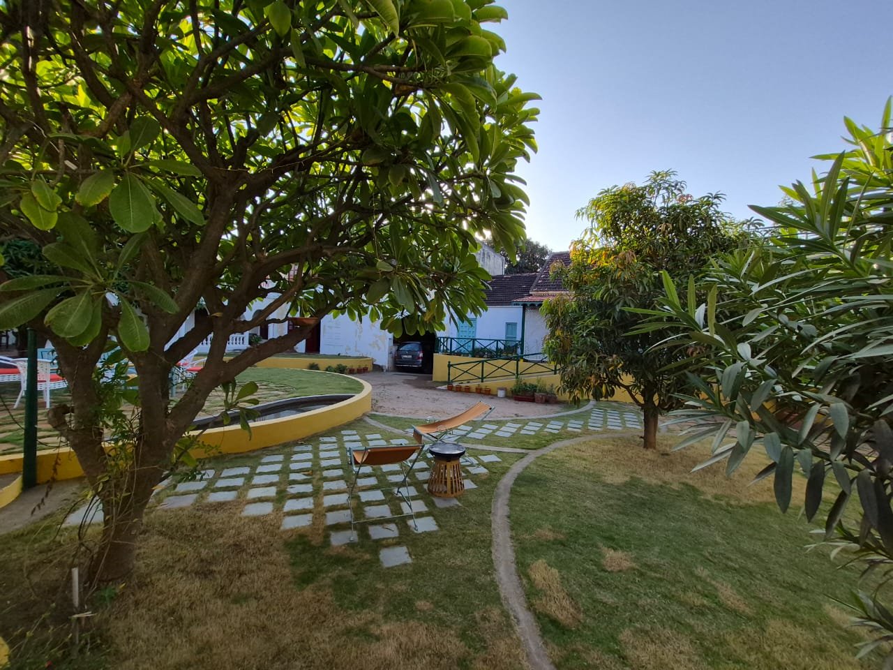 A peaceful backyard garden with lush green trees, a hammock, a small table, and chairs. A footpath curves through the yard, and a house with a car parked outside is visible in the background under a clear blue sky.