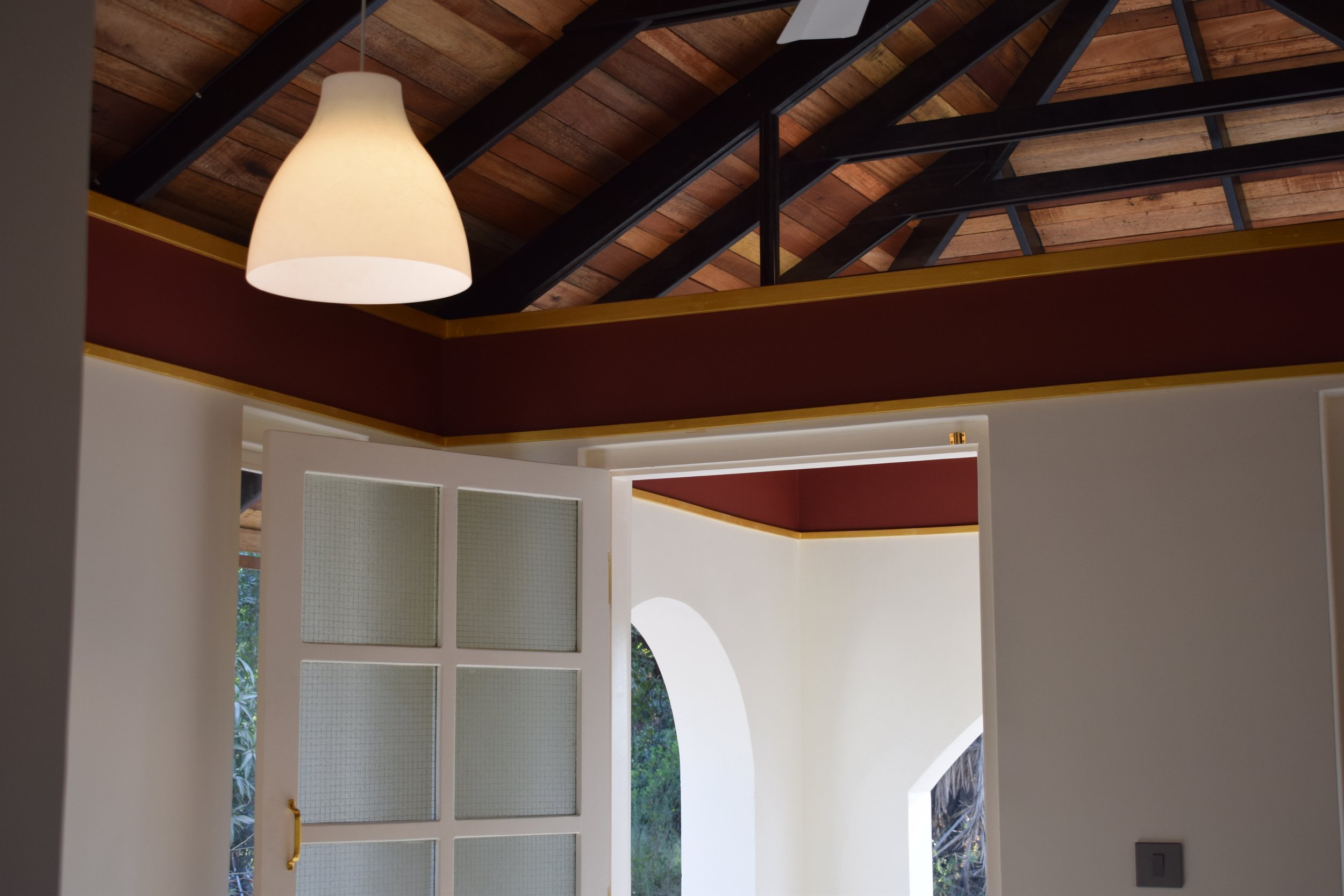 Interior view of a room with a high wooden ceiling, a white pendant lamp, and a white door with glass panels leading outside, showing greenery through the window.