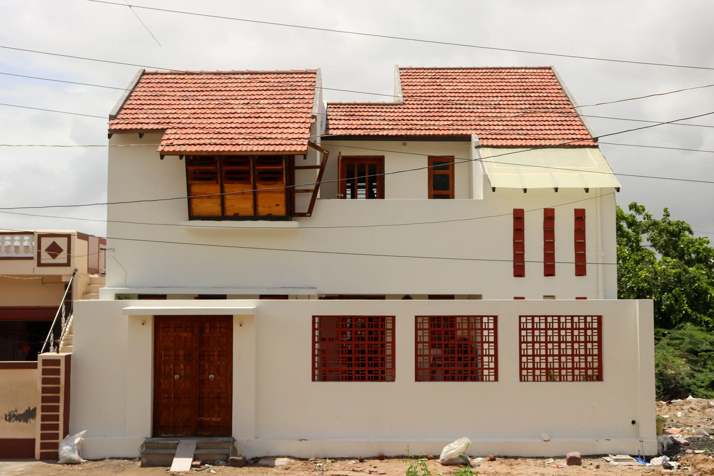 Modern two-story house with white exterior walls, wooden accents, and a red tiled roof, surrounded by a white perimeter wall with red decorative metalwork.