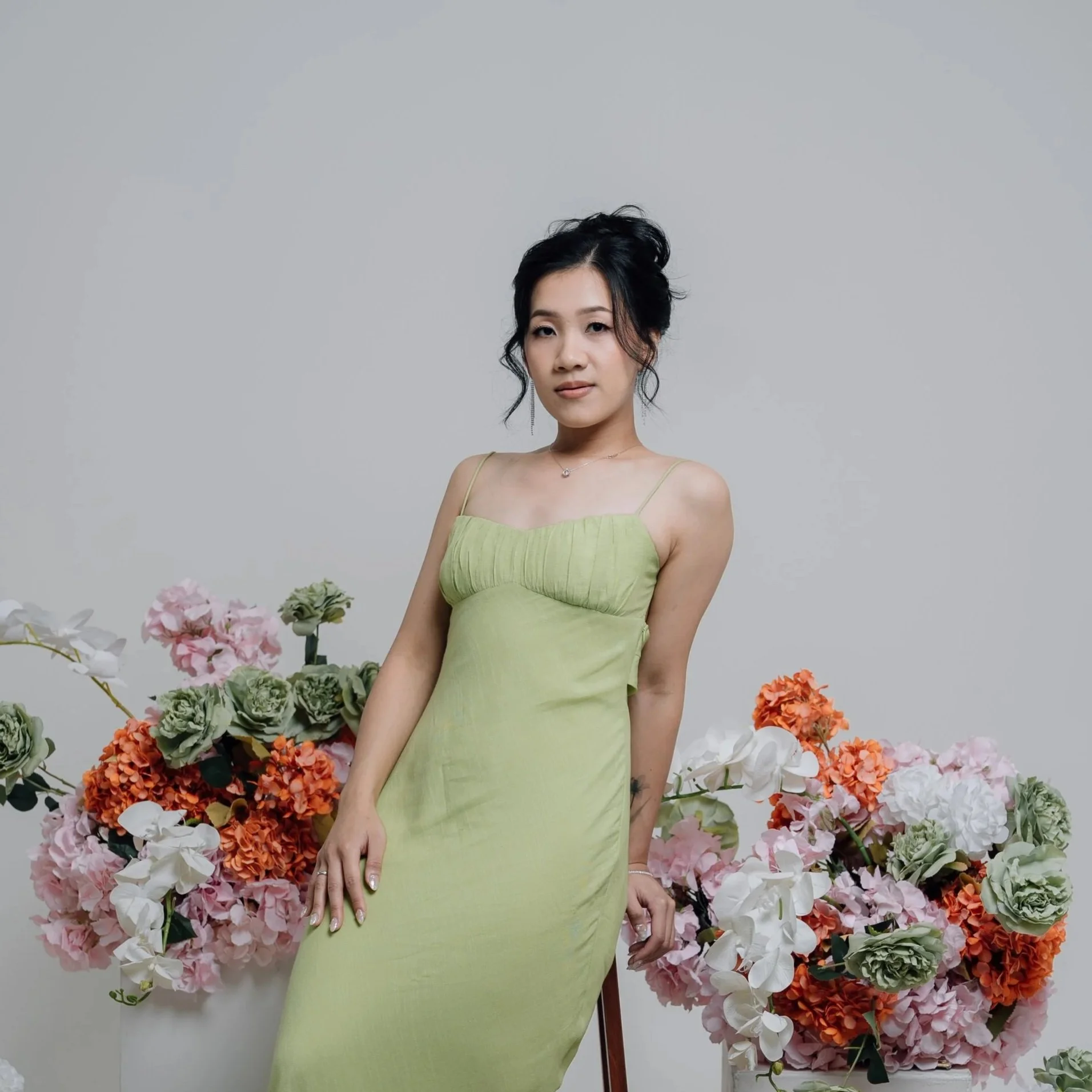 A woman with black hair styled in an elegant updo, wearing a light green spaghetti strap dress, posing in front of a plain light gray background with large, colorful flower arrangements including pink, orange, white, and green flowers.