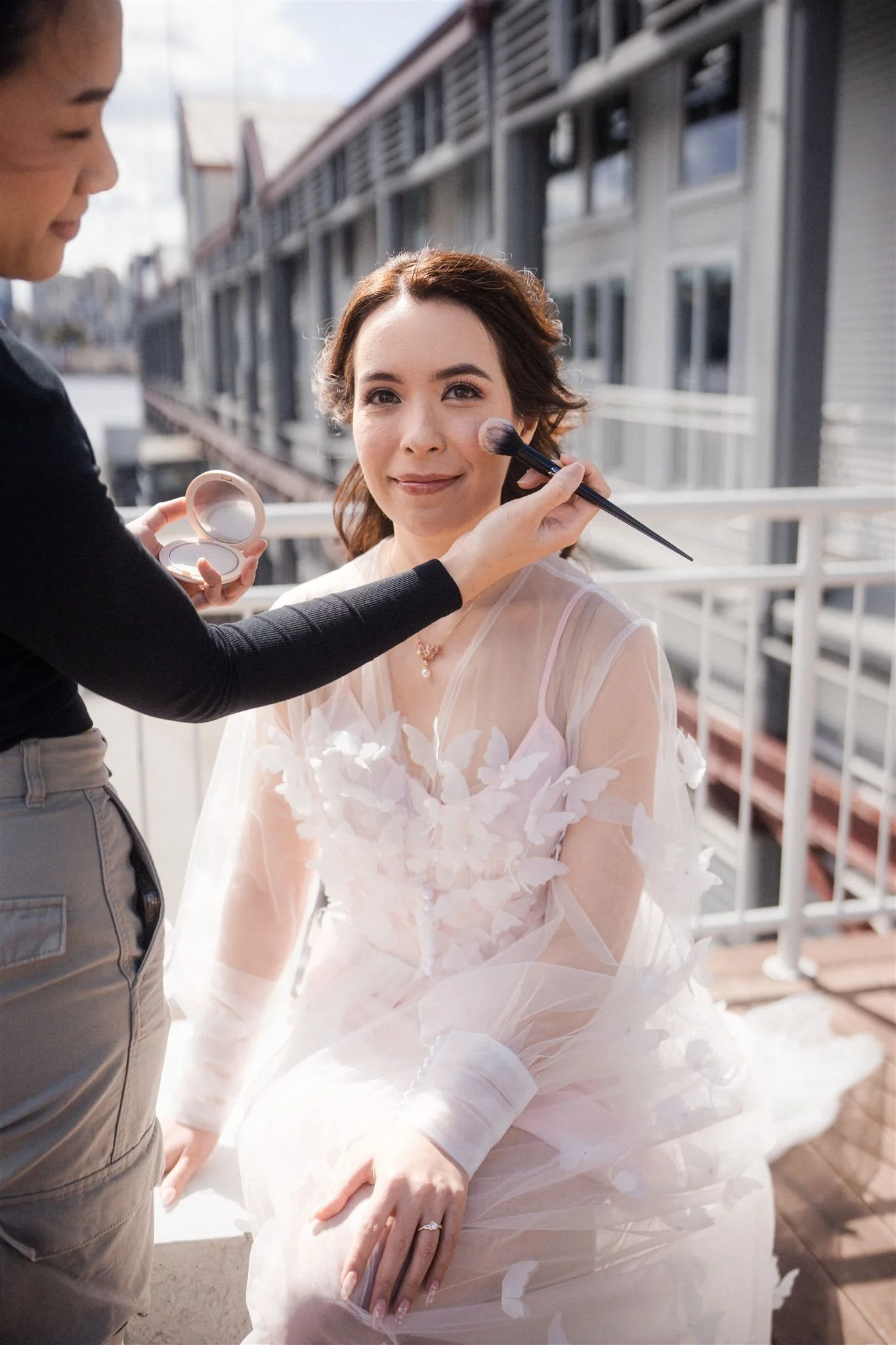 A woman in an elegant, light pink dress with floral details is having makeup applied outdoors by a makeup artist. The woman is smiling slightly and is sitting on a white surface with a cityscape in the background.