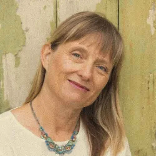 Veechi Stuart, the executive director of Varuna The National Writers' House sitting on a concrete step in front of a grey door, smiling at the camera. She is wearing a white top, brown pants, black sandals.