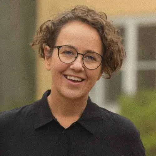 Amy Sambooke, the program advisor for The Blue Mountains Writer's Festival. She has curly hair, glasses, and a dark blue collared shirt, smiling outside Varuna The Nationals Writers' House. 