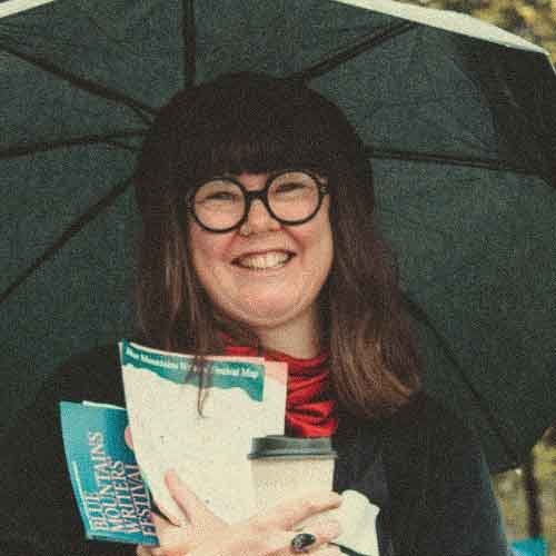 Erin English, the venue manager for the Blue Mouatins Writers' Festival. She has light brown hair and is smiling while sitting against a yellow wooden wall, wearing a colourful floral top and black pants.
