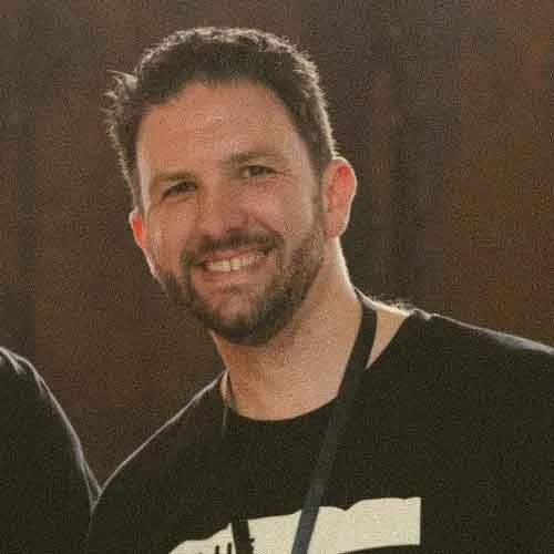 Chris Cannell, the communication and Marketing director at Varuna The National Writers' House. He is smiling and wearing a Black and white Blue Mountains Writers' Festival T-shirt and a lanyard, standing indoors.