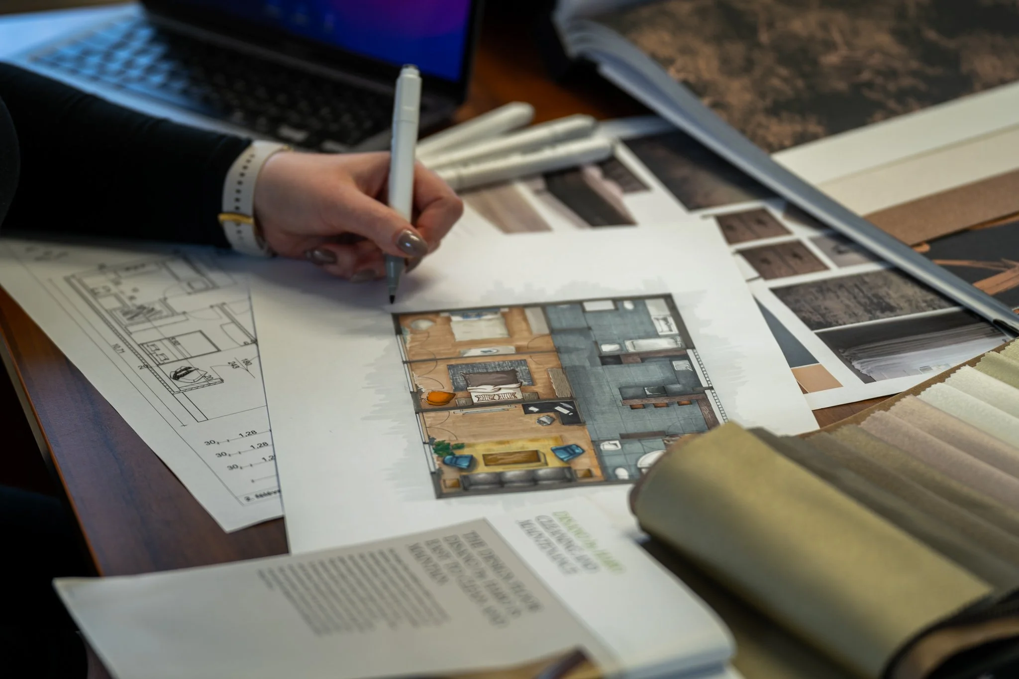 A person working on an interior design plan with architectural drawings, including a floor layout with furniture and color samples on a table.
