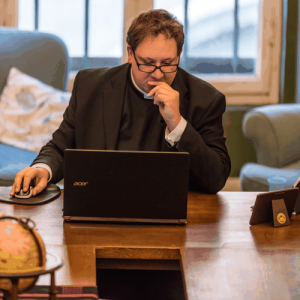 Man in a suit working on a laptop at a wooden table in a cozy room with large windows.