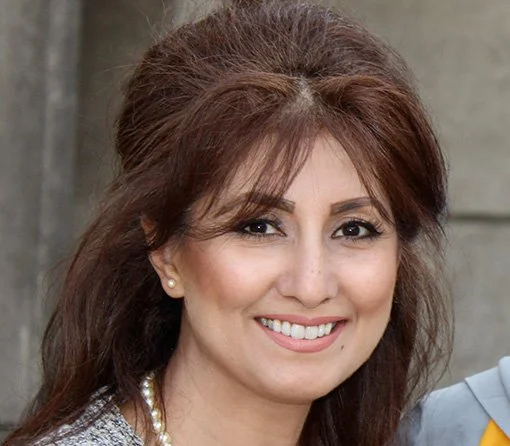 A smiling woman with long brown hair and pearl earrings, posing outdoors.
