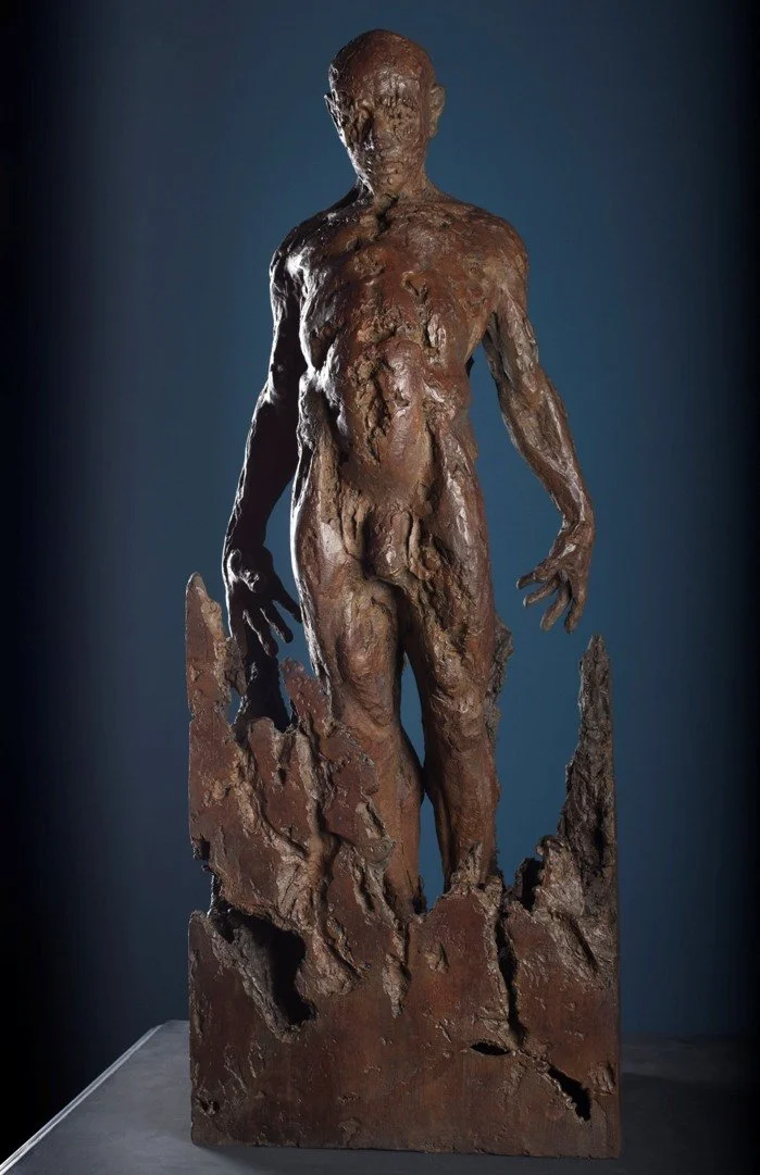 Bronze sculpture of a standing human figure with rough, textured surface, mounted on a jagged base.