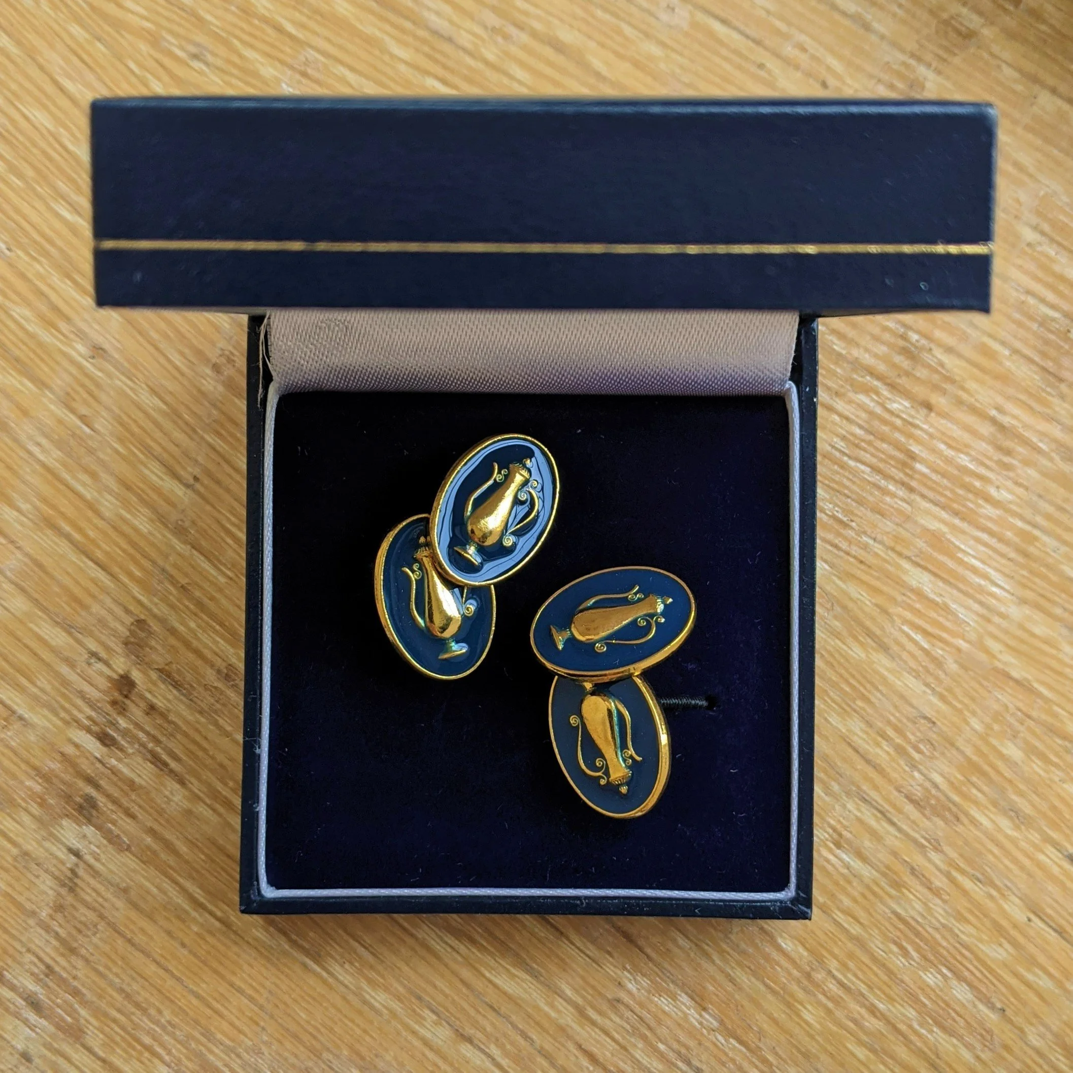 A pair of vintage-style earrings with blue oval bases and gold hummingbird designs inside, presented in a black jewelry box with a beige interior on a wooden surface.