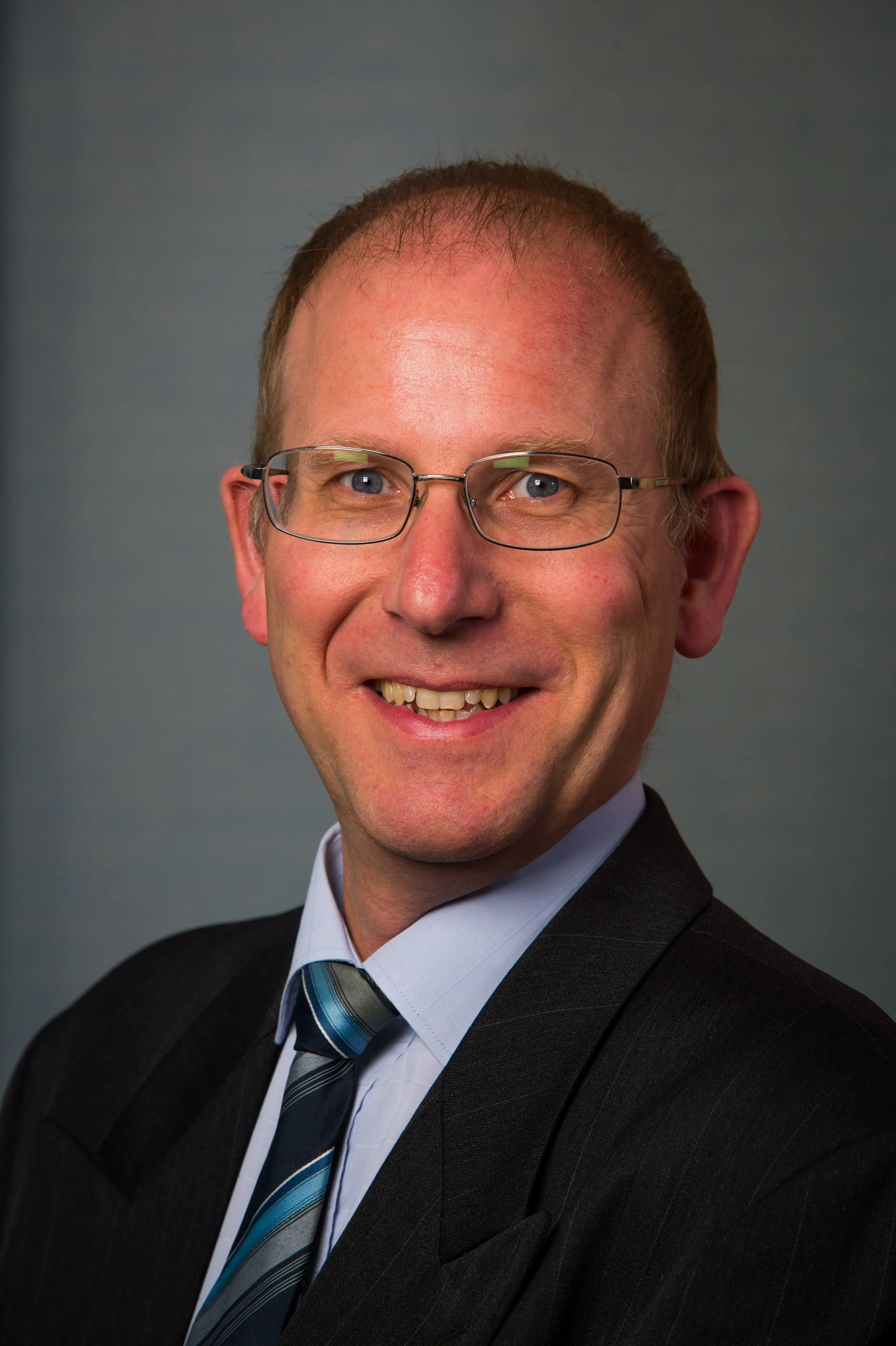 Professional headshot of a smiling man with glasses, wearing a dark suit, light blue shirt, and a striped tie, against a neutral gray background.
