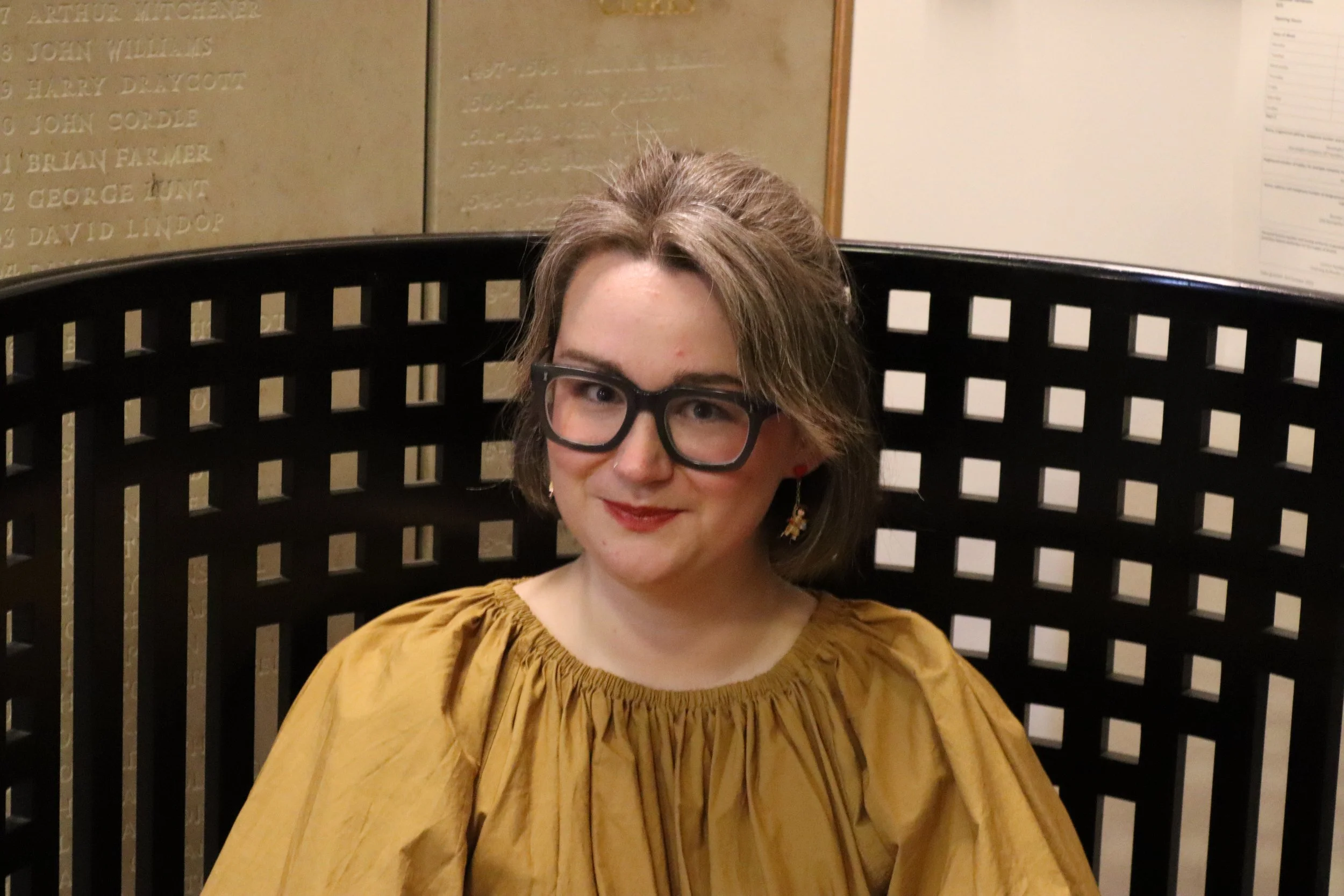 Young woman with glasses smiling at camera in a formal dining setting, surrounded by other people dressed in formal attire, with paintings and shields displayed on the red wall behind her.