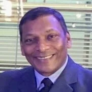 A man smiling in a professional setting, wearing a suit and tie with a background of window blinds.