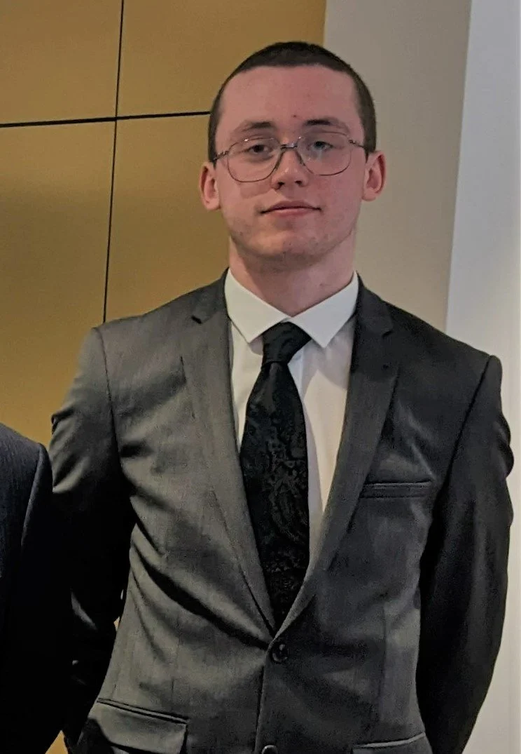 A young man wearing glasses dressed in a gray suit, white shirt, and black tie, standing in front of a wooden-paneled wall.