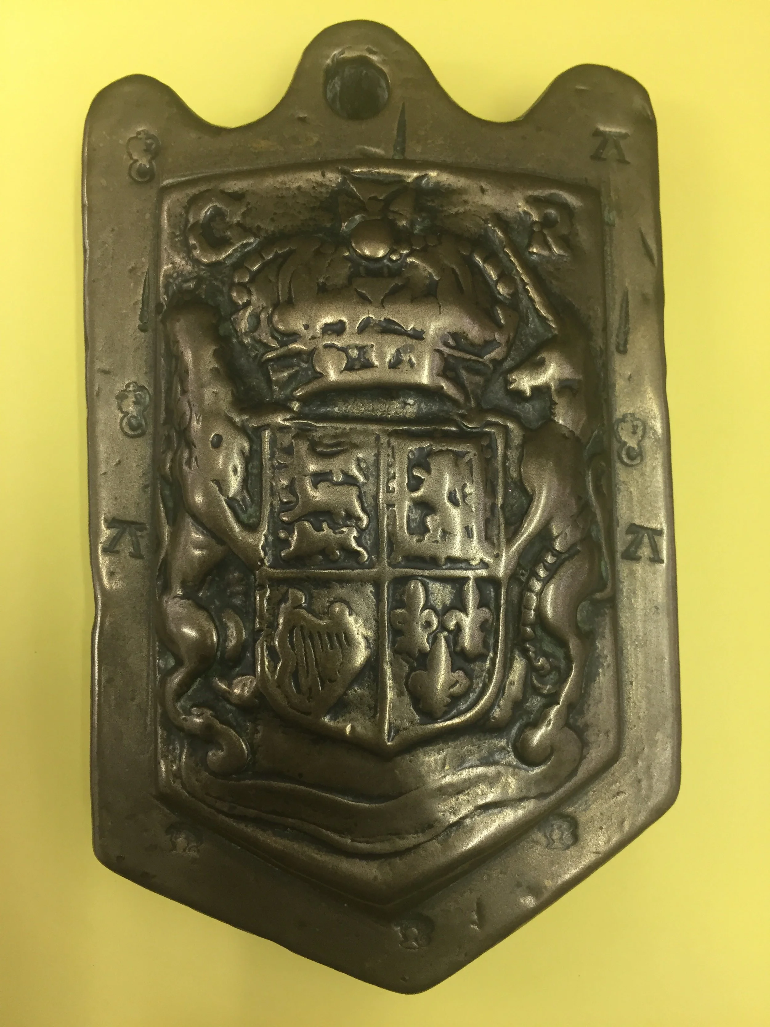 A metal badge or plaque featuring a coat of arms with a crowned lion and a unicorn, divided into four quadrants with various heraldic symbols, mounted on a yellow background.
