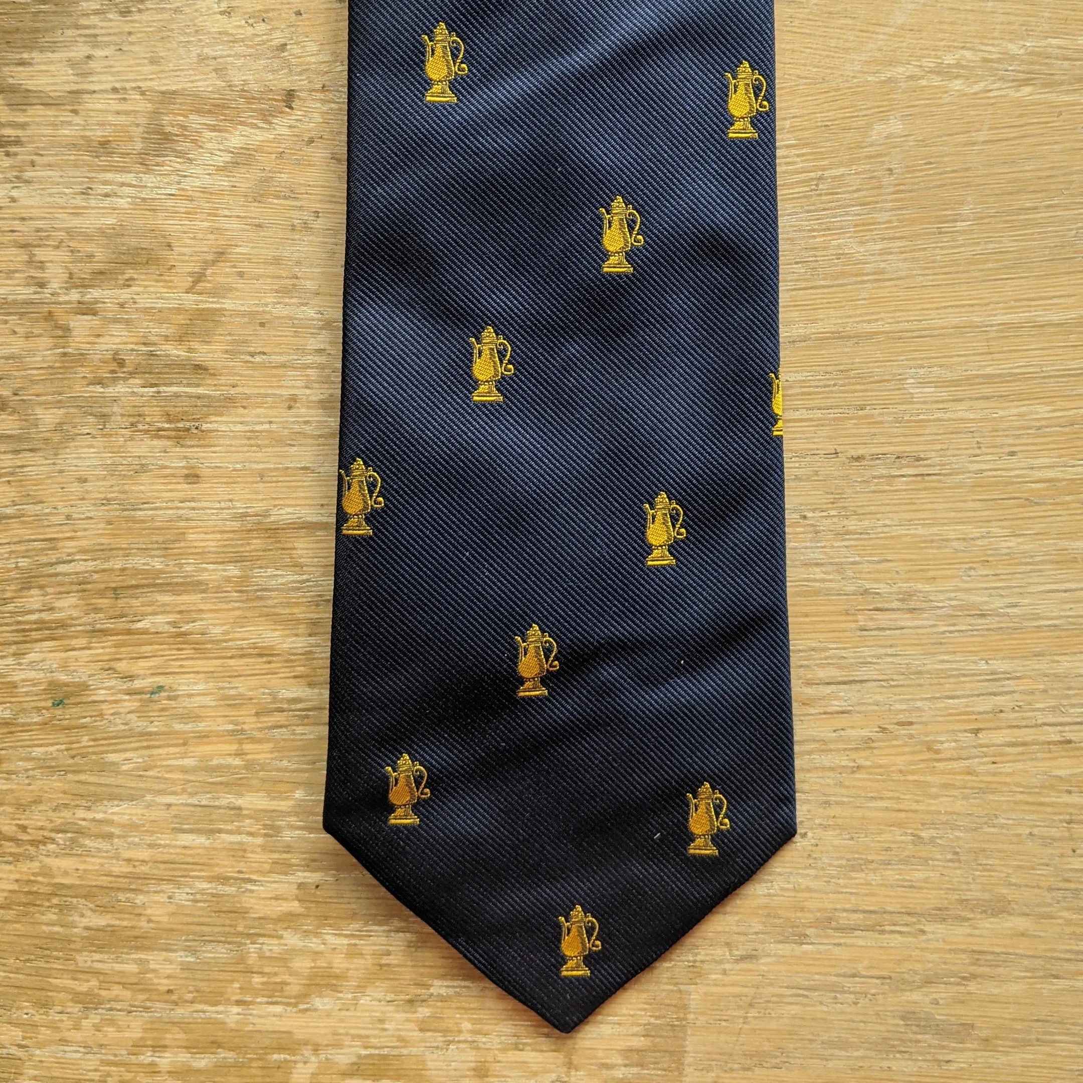 A dark blue tie with small gold patterns of a trophy or urn, laid flat on a wooden surface.