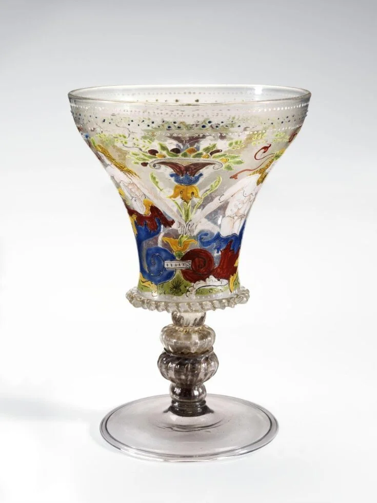 A decorative glass goblet with a painted floral and abstract design on the bowl, featuring colors like red, blue, green, and yellow, with a clear, textured stem and base.