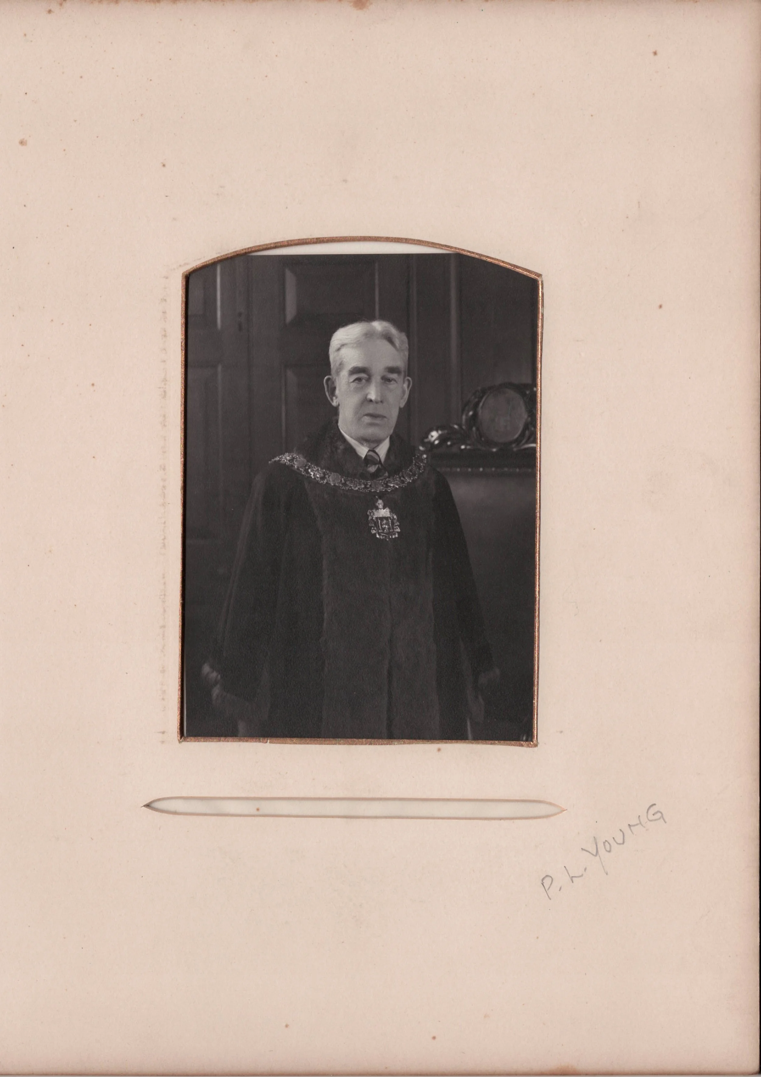 Black and white portrait of an older man in formal academic robes, standing inside a room with wood-paneled walls, framed by a mat with a handwritten signature "P. H. Young".