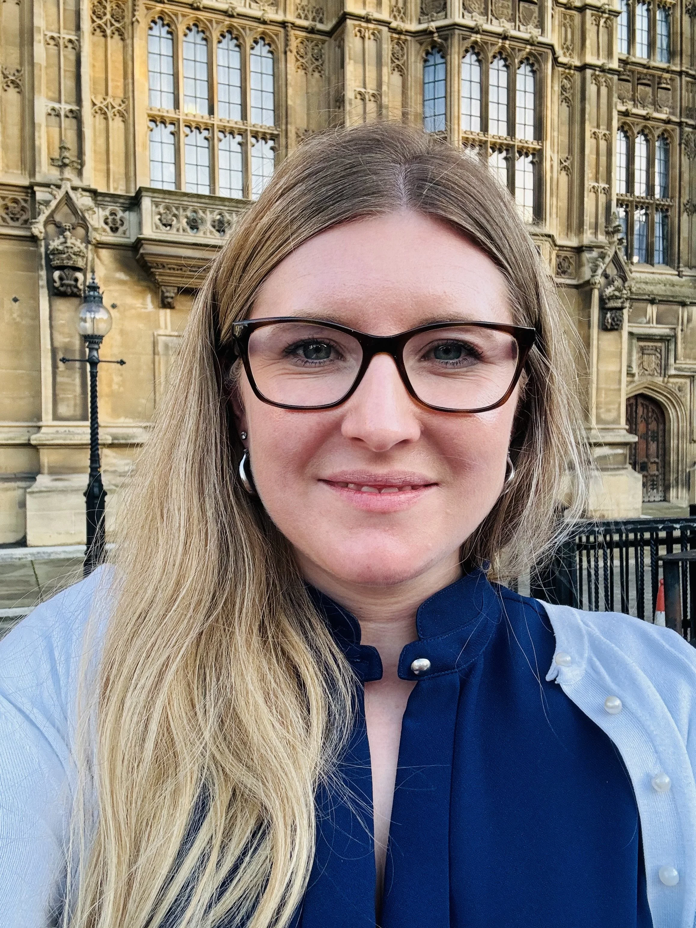 A woman with long blonde hair, glasses, and silver earrings taking a selfie in front of a historic building with ornate architecture.