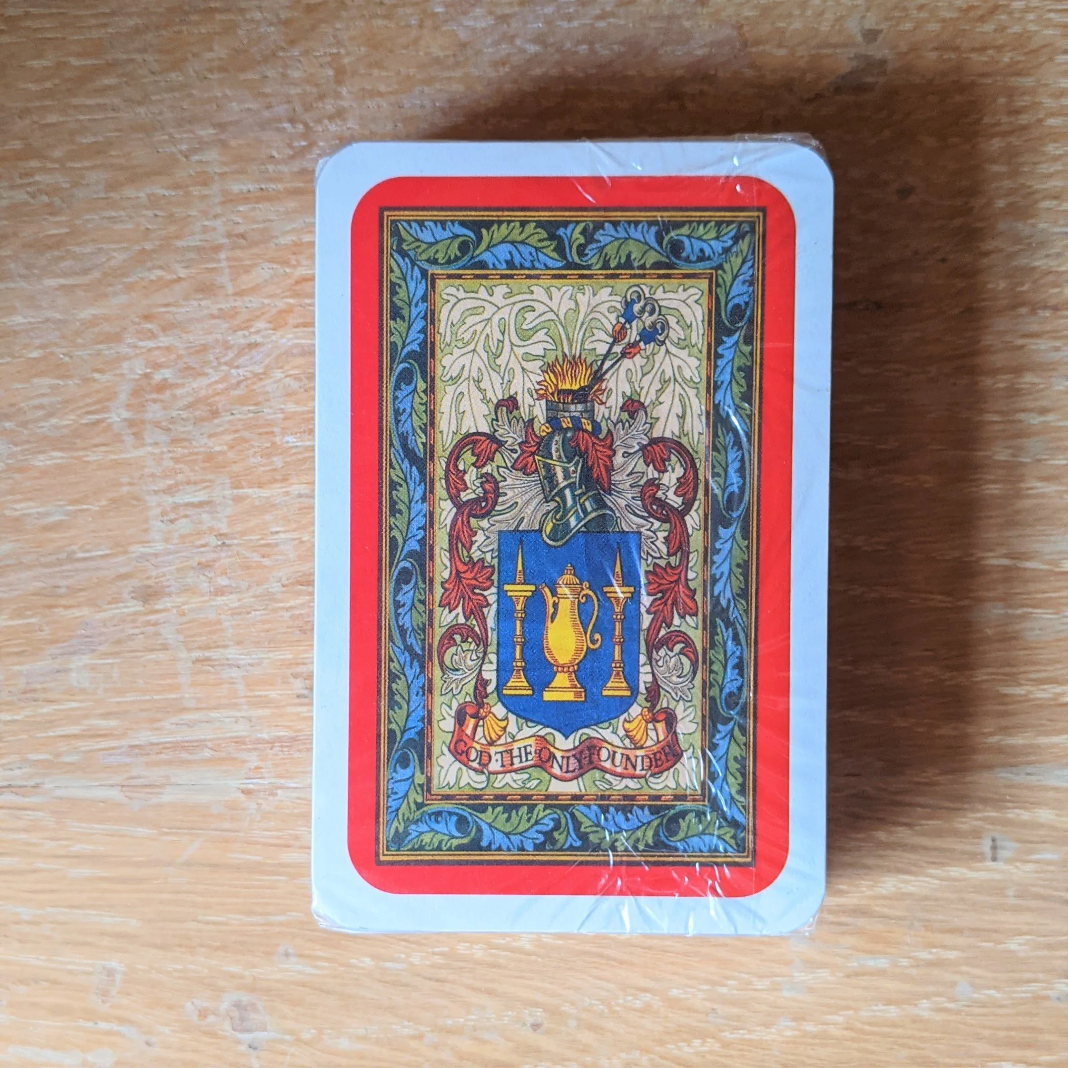 Back of a playing card with a colorful medieval crest featuring a blue shield, golden candlesticks and a teapot, framed with intricate red, green, and blue patterns.