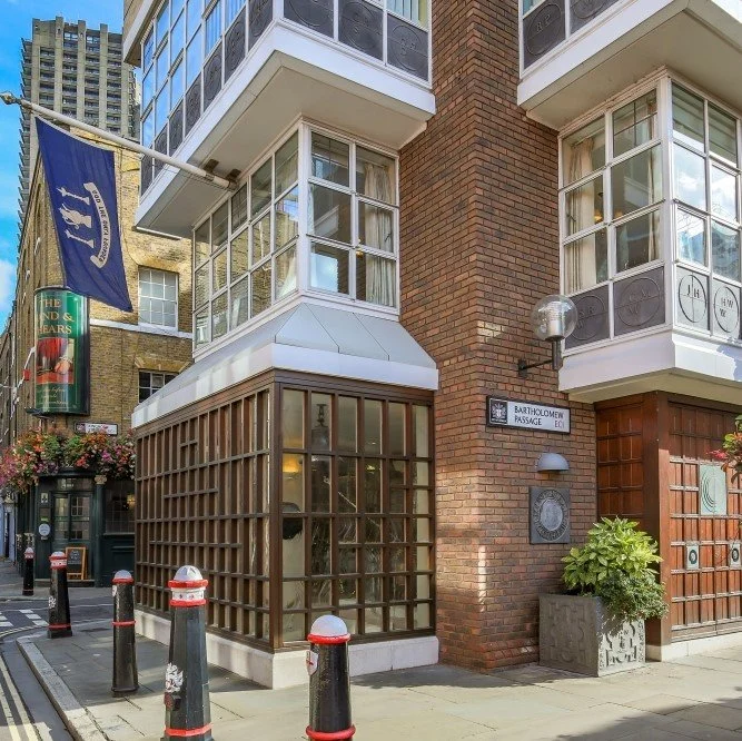 Corner of a brick building on a city street with a sign that says Bartholomew Passage, a lamp, a potted plant, and a glass-enclosed corner ice cream shop or cafe with large windows and metal framing