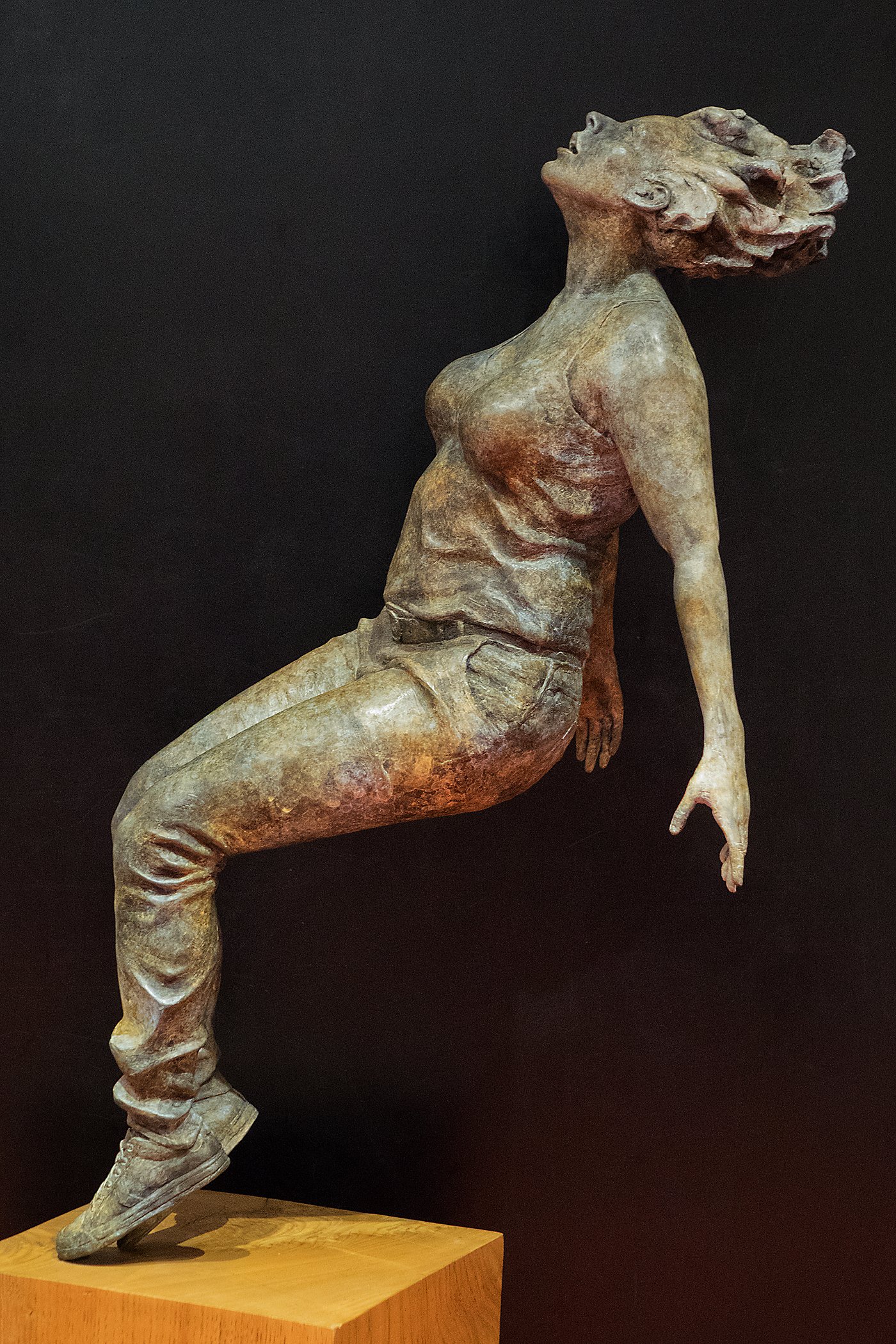 Bronze sculpture of a woman with hair flowing backward and her body in a reclining position on a wooden base, set against a black background.