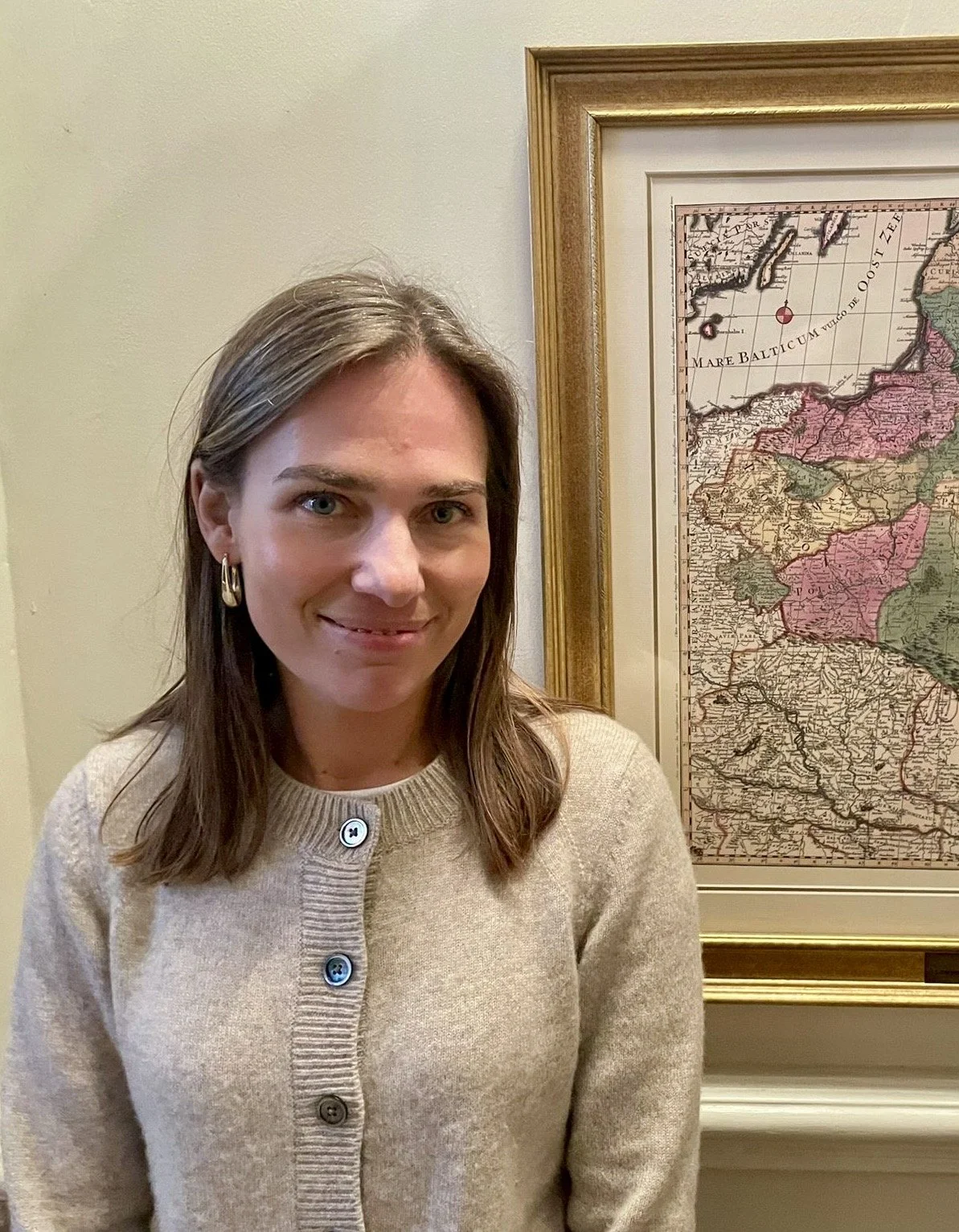 A woman with shoulder-length brown hair and earrings standing in front of a framed map of Asia.