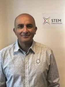 A man standing indoors, wearing a white patterned dress shirt, smiling at the camera. Behind him is a white wall with a logo that reads 'STEM Ambassadors'.