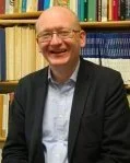 Smiling man in glasses wearing a suit jacket and light blue shirt, standing in front of bookshelves.