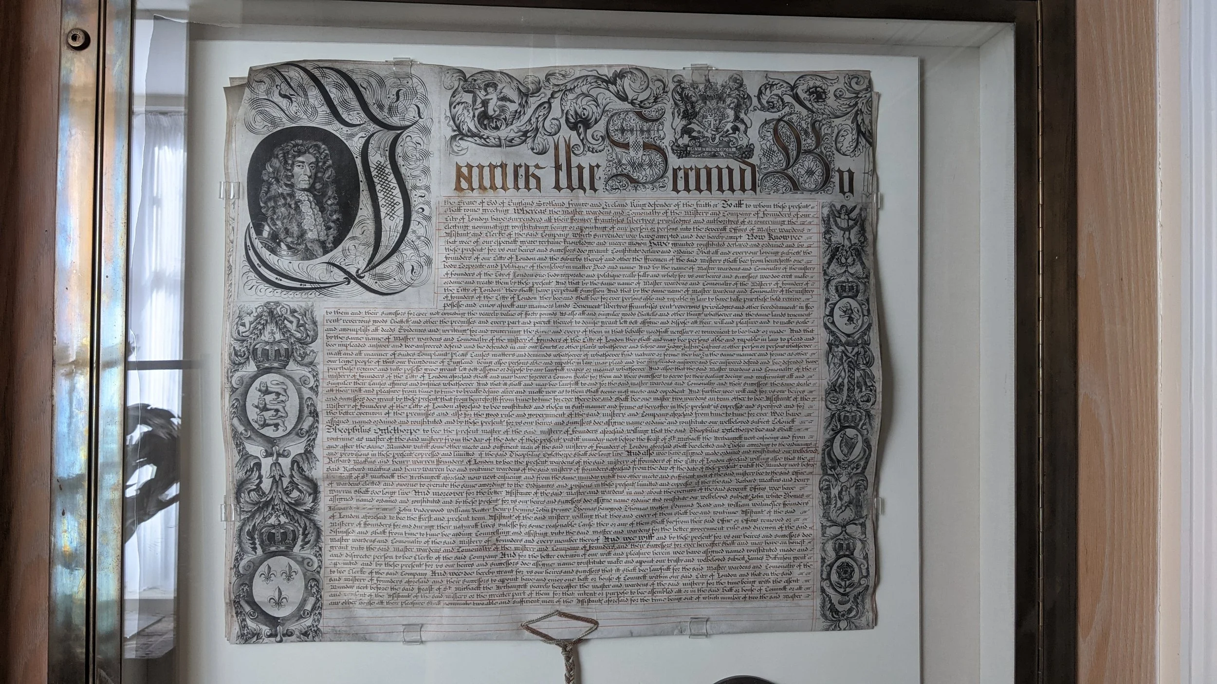 An antique parchment document framed behind glass, featuring ornate calligraphy, illustrated coats of arms, and decorative borders with lion and fleur-de-lis symbols.