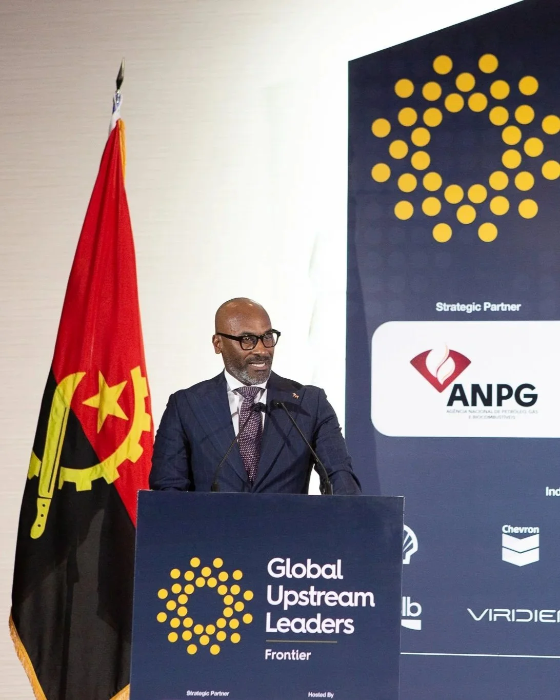 A man in a suit and glasses standing at a podium with a microphone, delivering a speech at a conference called 'Global Upstream Leaders.' Behind him, a large screen displays logos and text, including 'Strategic Partner' and the ANPG logo. To his left, a red flag with yellow and black symbols is visible.