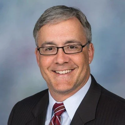 A man in a suit with glasses and a red tie with a blur background.