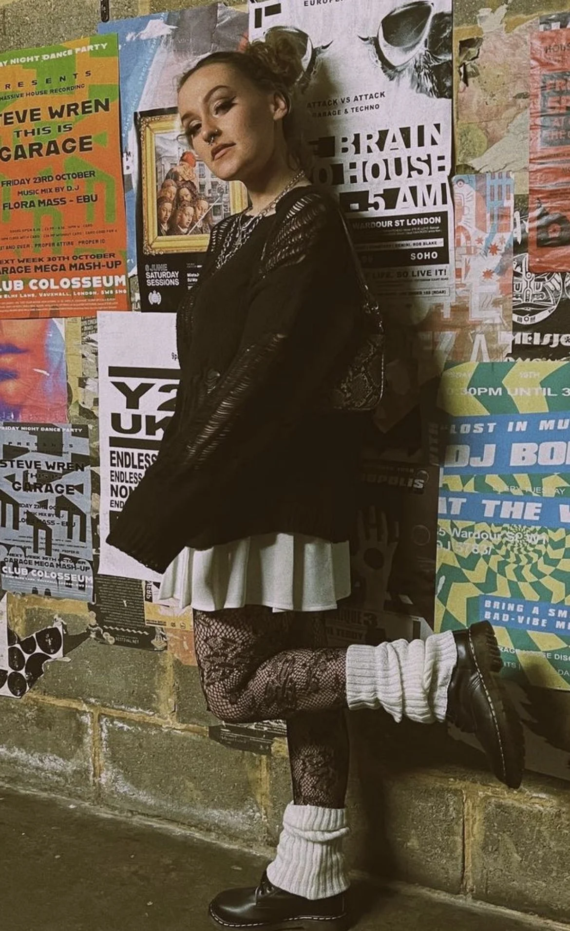 Person with makeup, curly hair in a bun, and gothic style clothing posing in front of a wall covered with colorful posters.