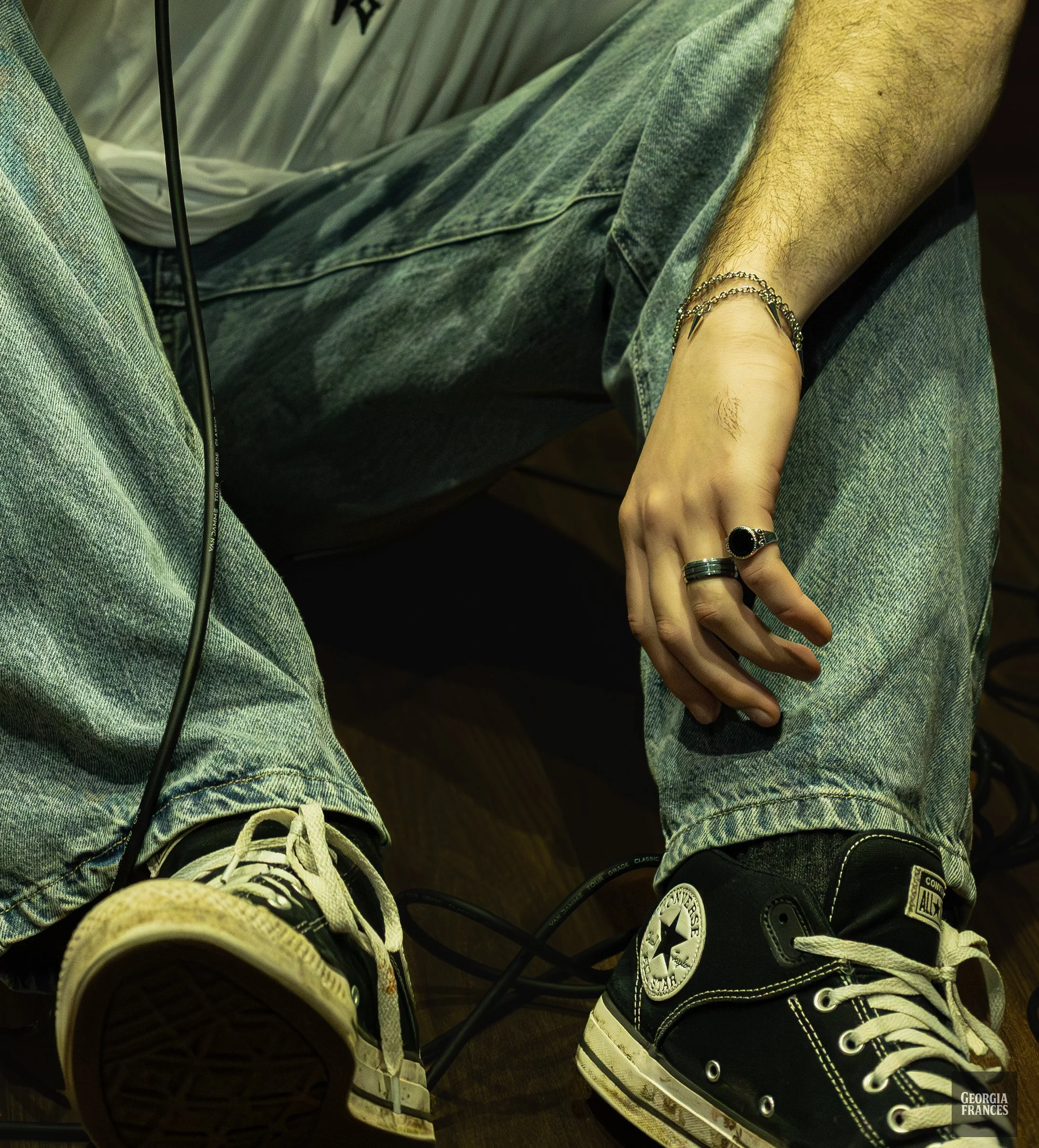 Close-up overhead view of RUoK wearing blue jeans and Converse sneakers, with one hand displaying rings and bracelets.