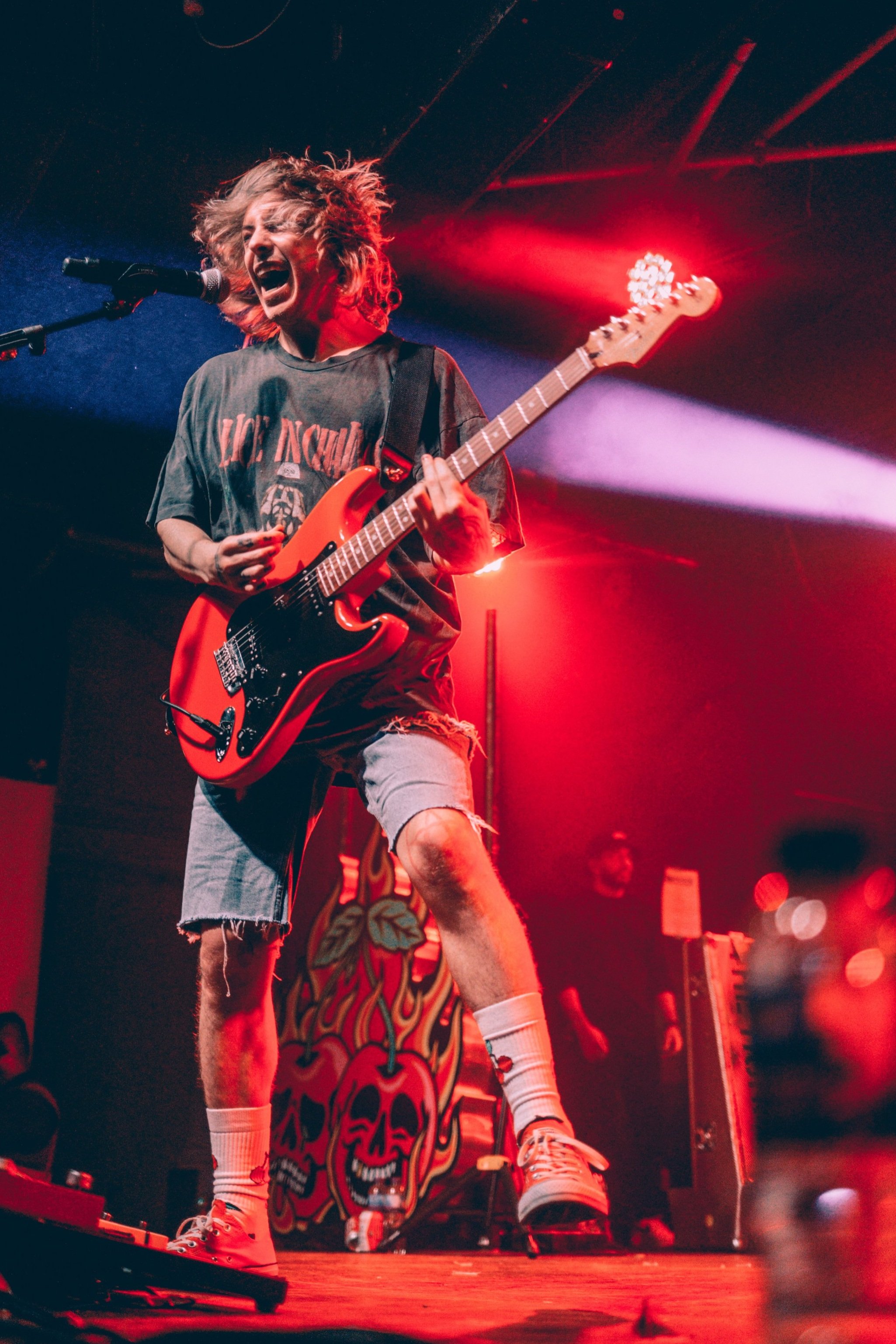 Ryan from The Hunna singing into a microphone while playing an electric guitar on a red-lit stage at the Trinity Centre, Bristol.