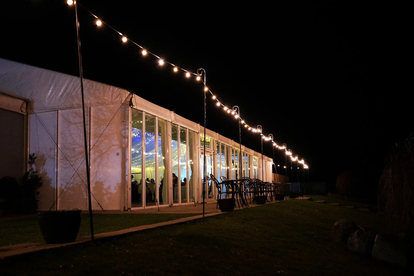 Nighttime scene of a large white event tent illuminated by string lights, with visible glass walls revealing a decorated interior with people inside, and outdoor tables and chairs set up nearby.