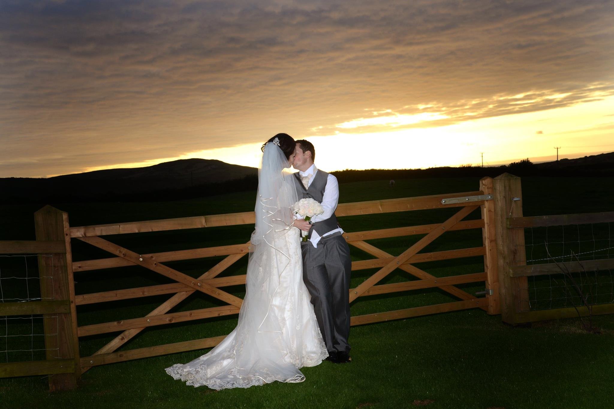 A bride and groom sharing a kiss outside at sunset, with the bride wearing a white wedding dress and veil, and the groom in a gray suit, holding a bouquet, standing by a wooden fence with a rural landscape in the background.