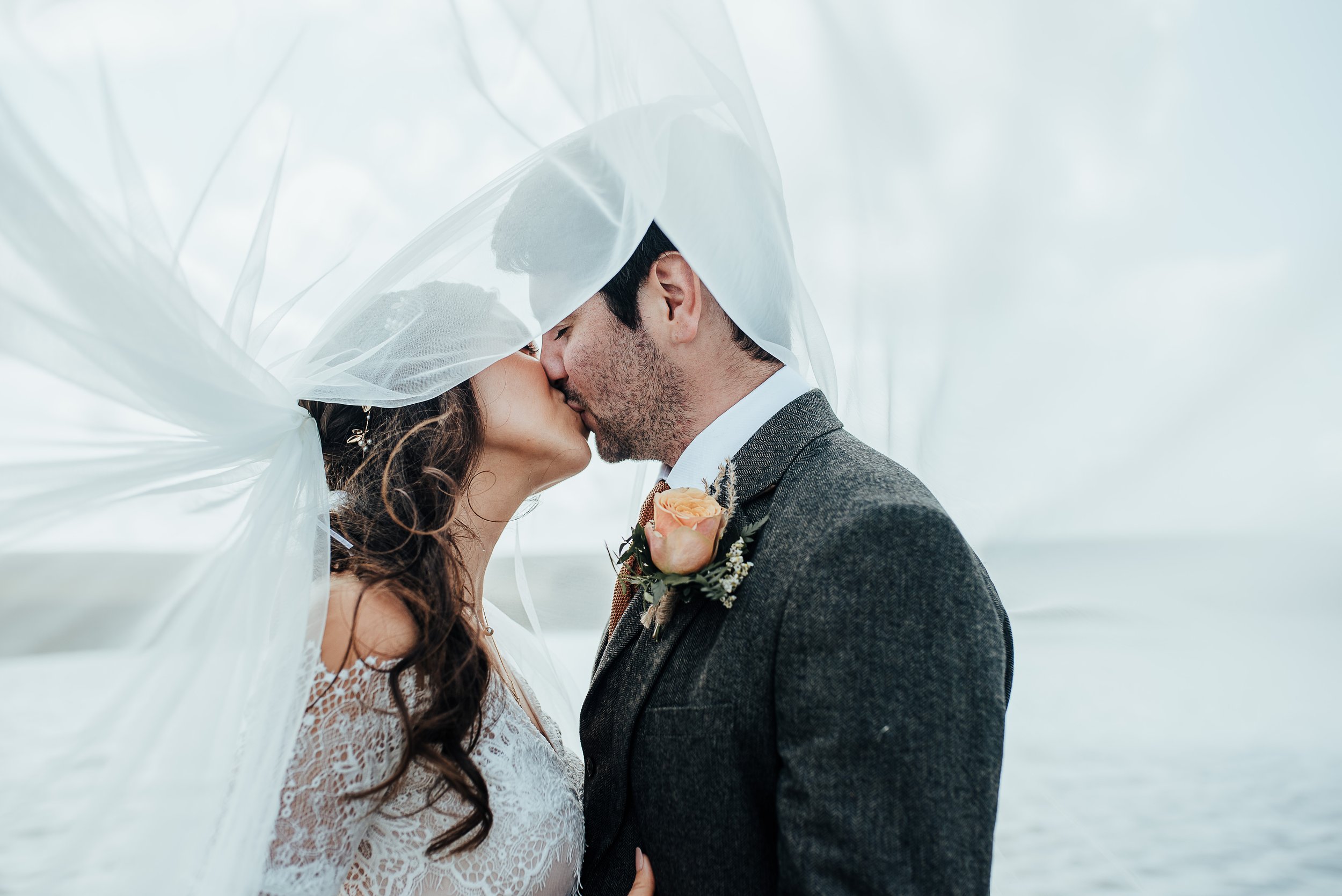 A bride and groom kiss outdoors, with the bride wearing a large white veil and a lace wedding dress, and the groom in a gray suit with a peach-colored rose boutonniere.