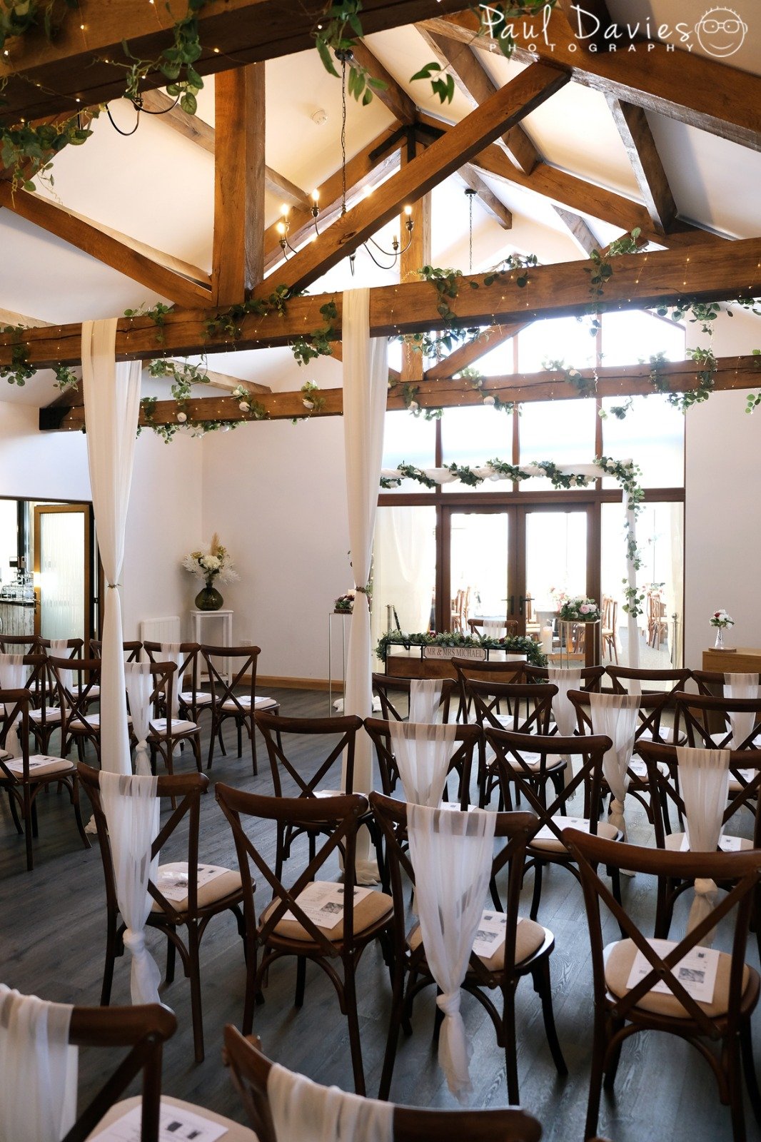 Indoor wedding ceremony setup with wooden chairs draped with white fabric ribbons, floral decorations on the ceiling beams and around the altar, and large windows allowing natural light in.