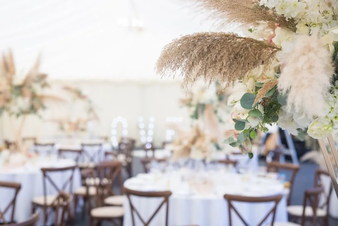 Elegant wedding reception setup with round tables, white tablecloths, wooden chairs, and floral centerpieces featuring pampas grass and greenery.