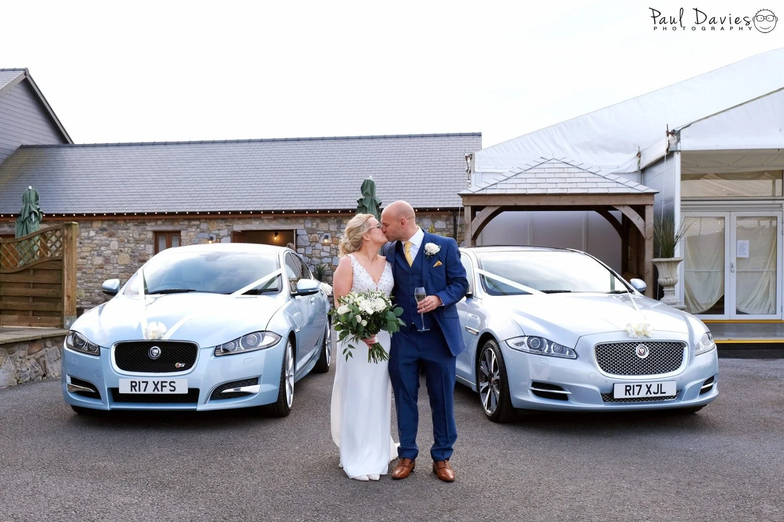 A bride and groom sharing a kiss in front of two silver luxury cars decorated with white ribbons at a wedding venue.